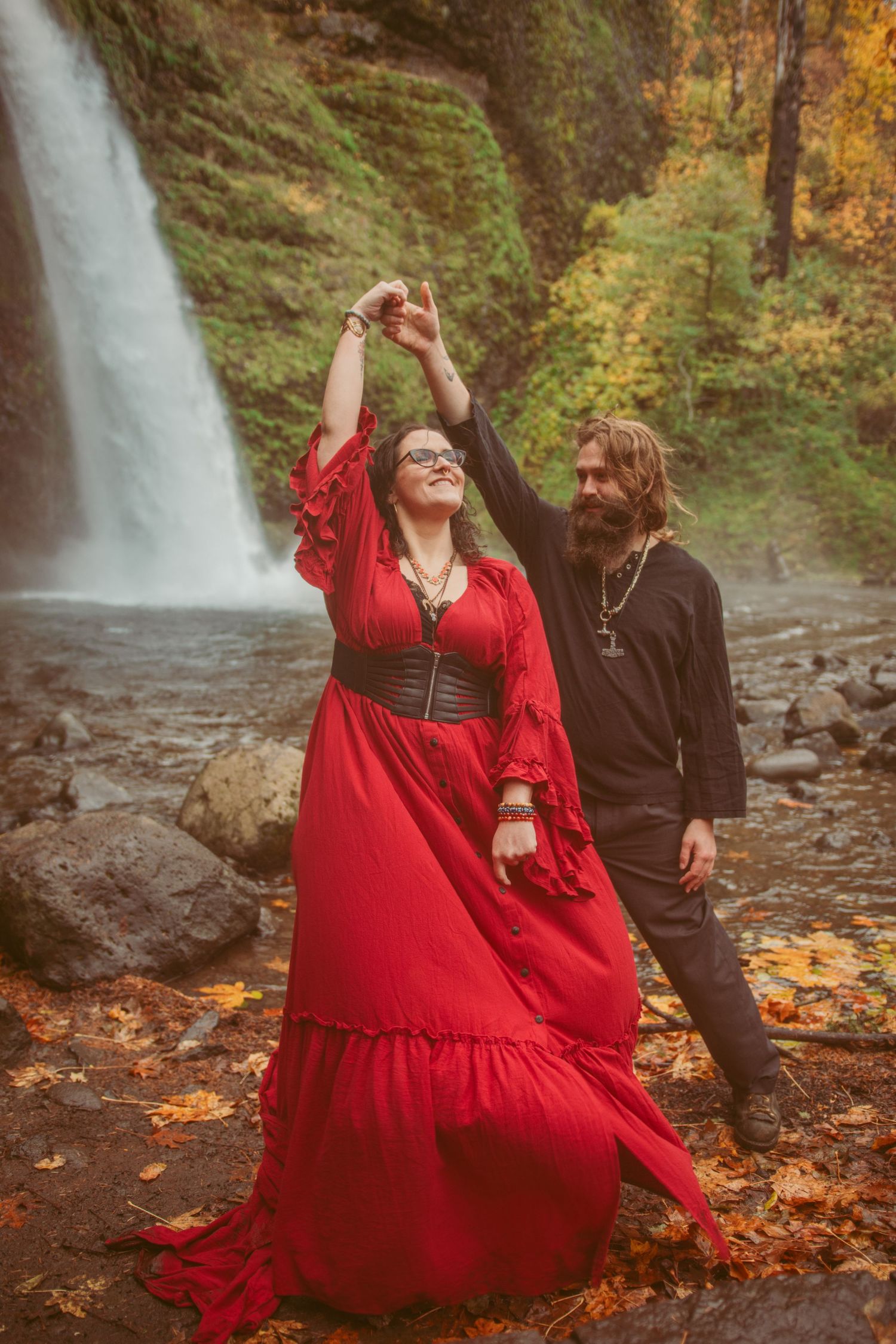 Dancing couple in red dress and black attire twirl by autumn waterfall.