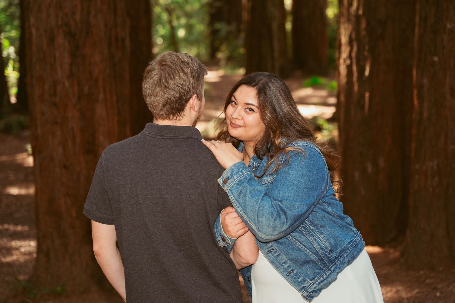 A tender moment between a couple in denim and casual wear against a backdrop of tall forest trees.