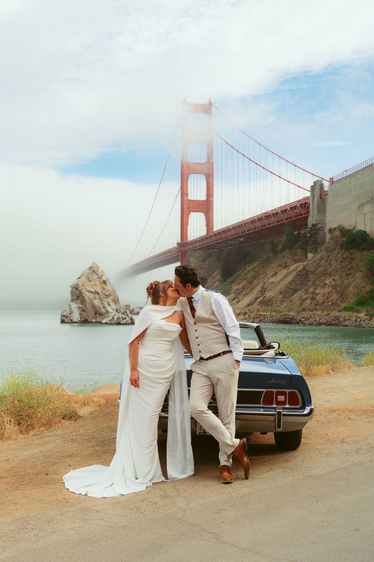 Bride and groom kissing by vintage car with Golden Gate Bridge backdrop on their wedding day.