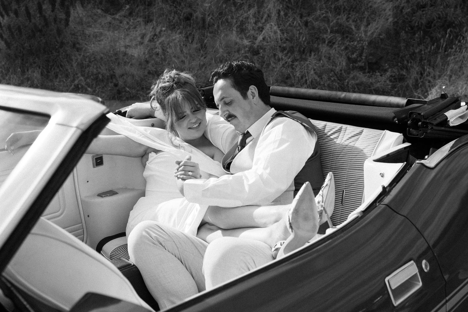 Black and white photo of a romantic couple cuddling in a convertible car outdoors.