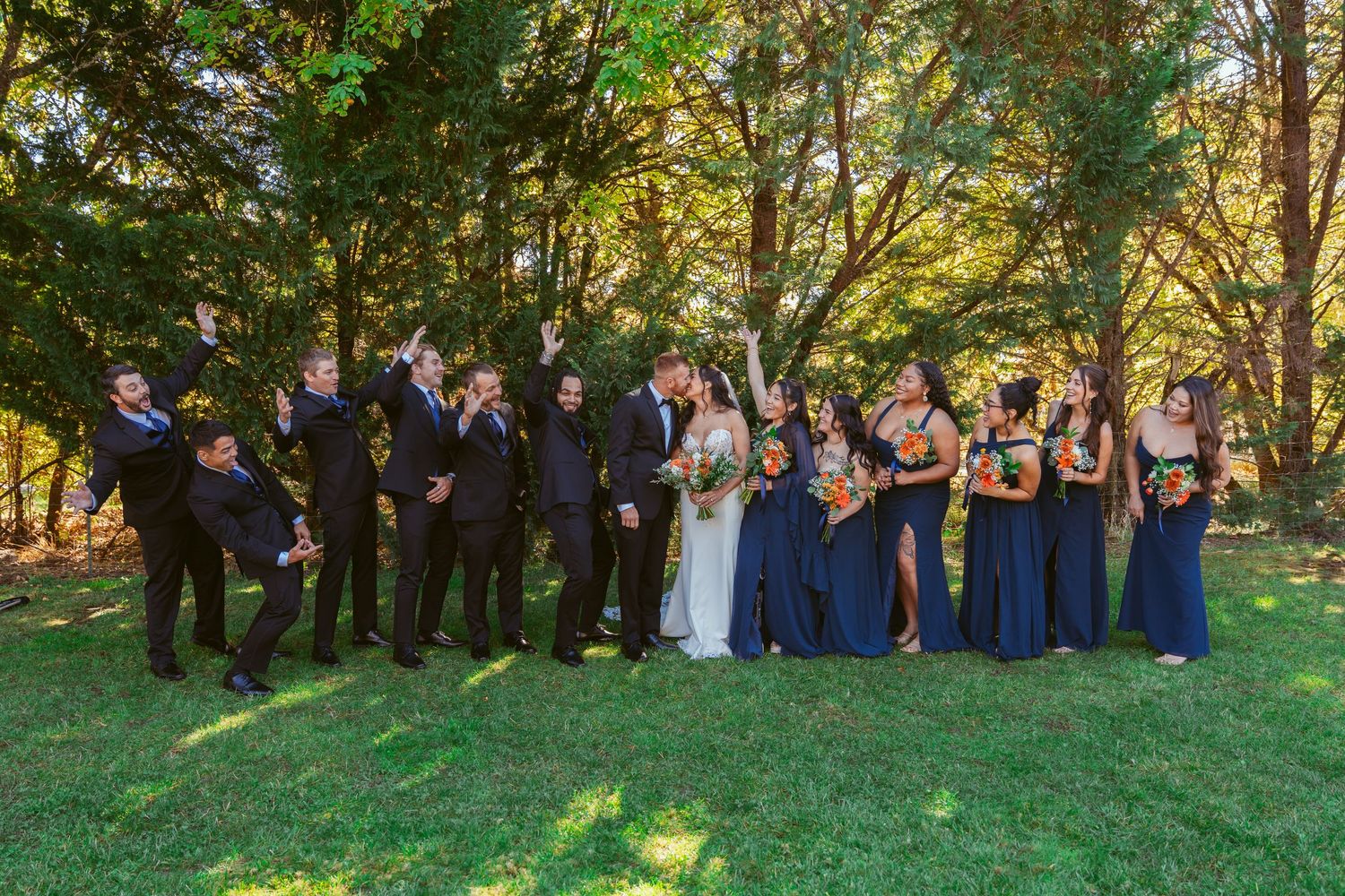 Full wedding party poses on lawn with bridesmaids in navy and groomsmen in black suits.