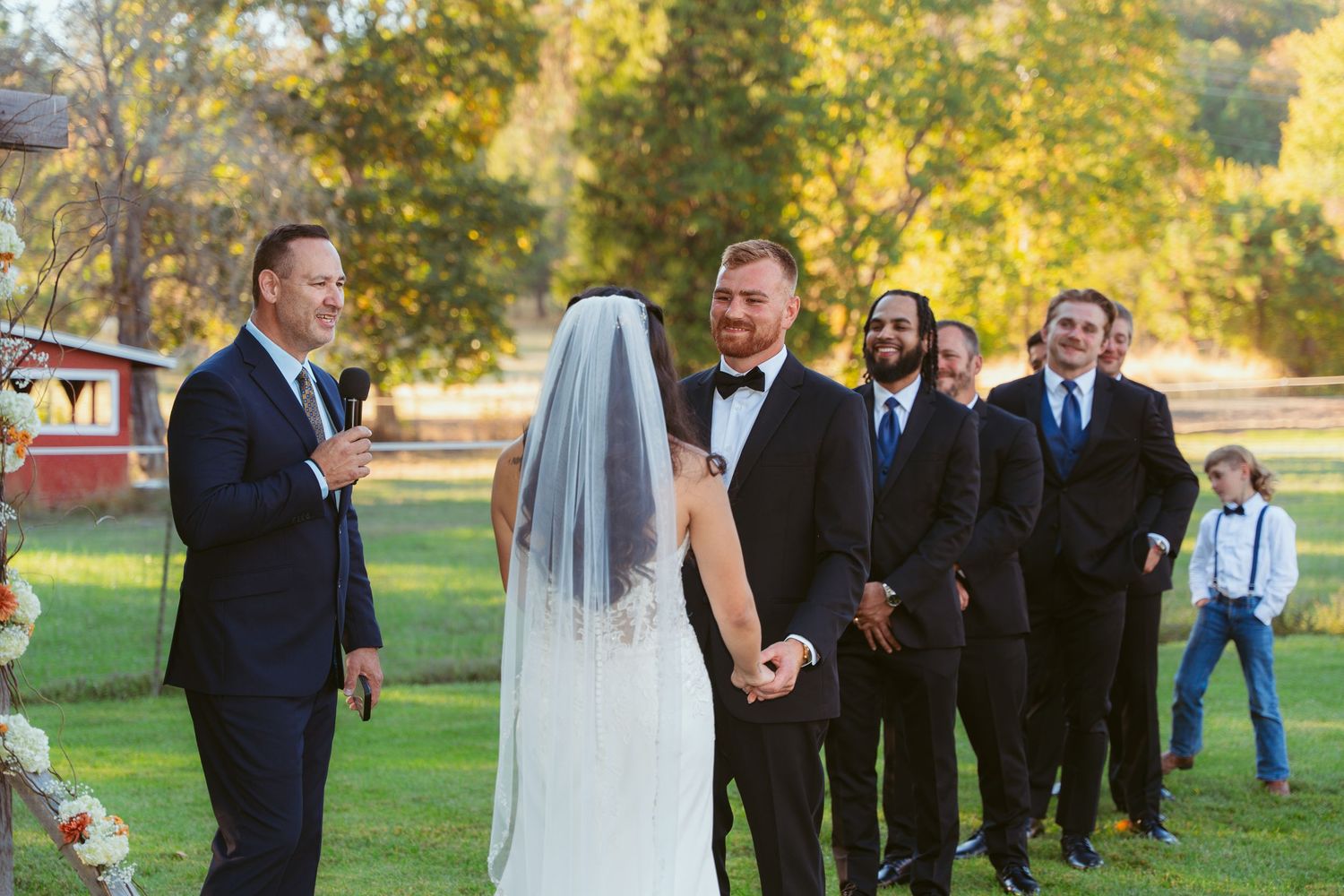 Outdoor wedding ceremony with attendees gathered on grassy lawn surrounded by fall trees.