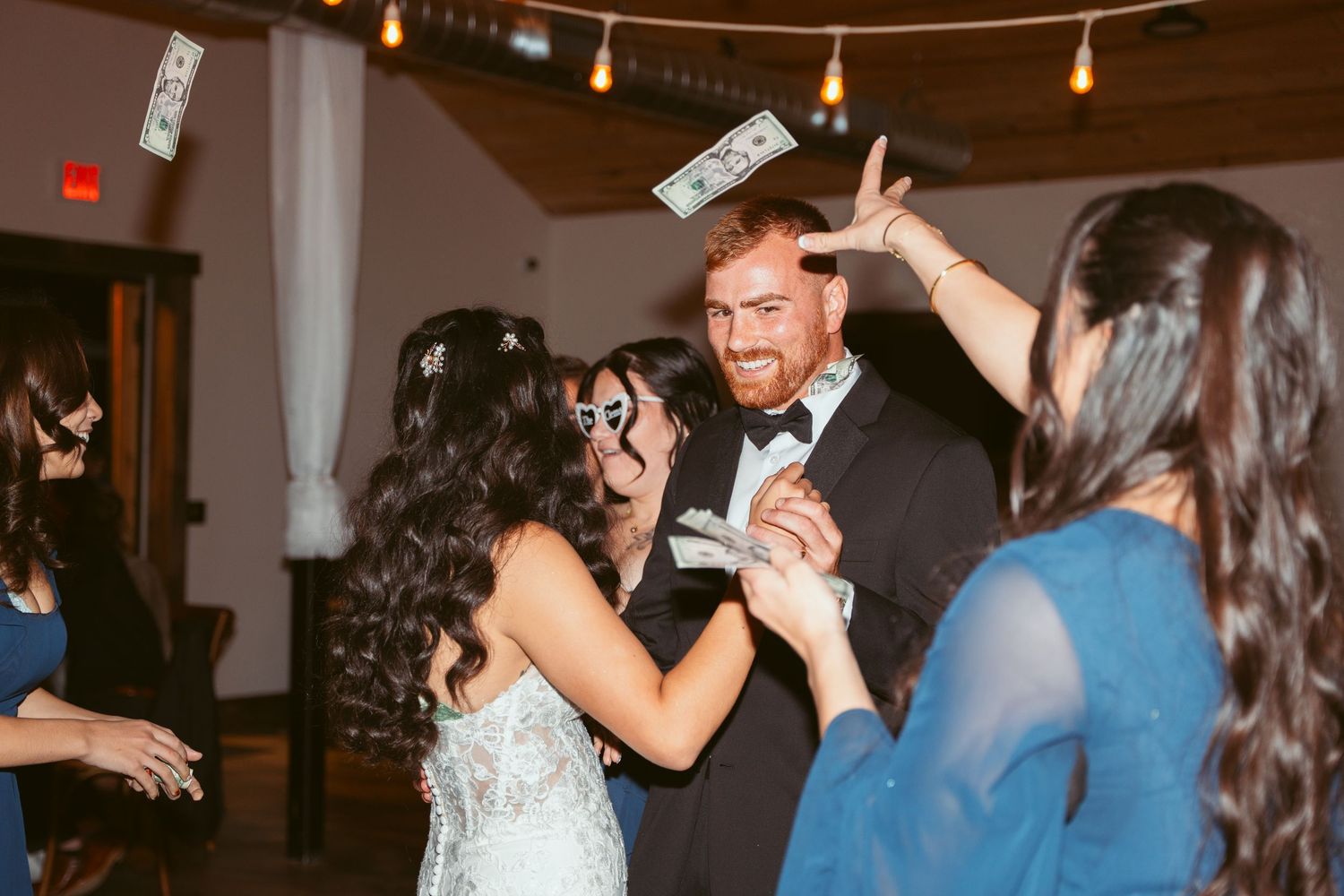 Couples dancing and celebrating at an indoor wedding reception with string lights overhead.