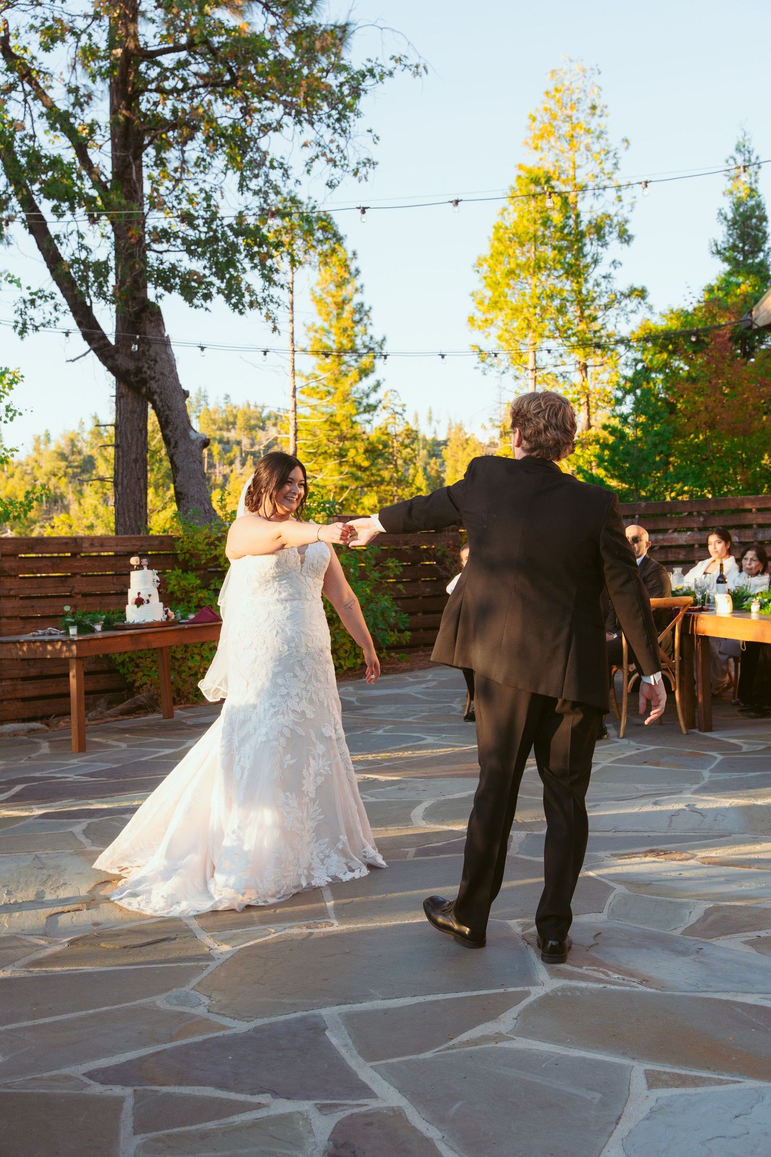 Newlyweds share a dance on outdoor stone patio surrounded by pine trees at sunset.