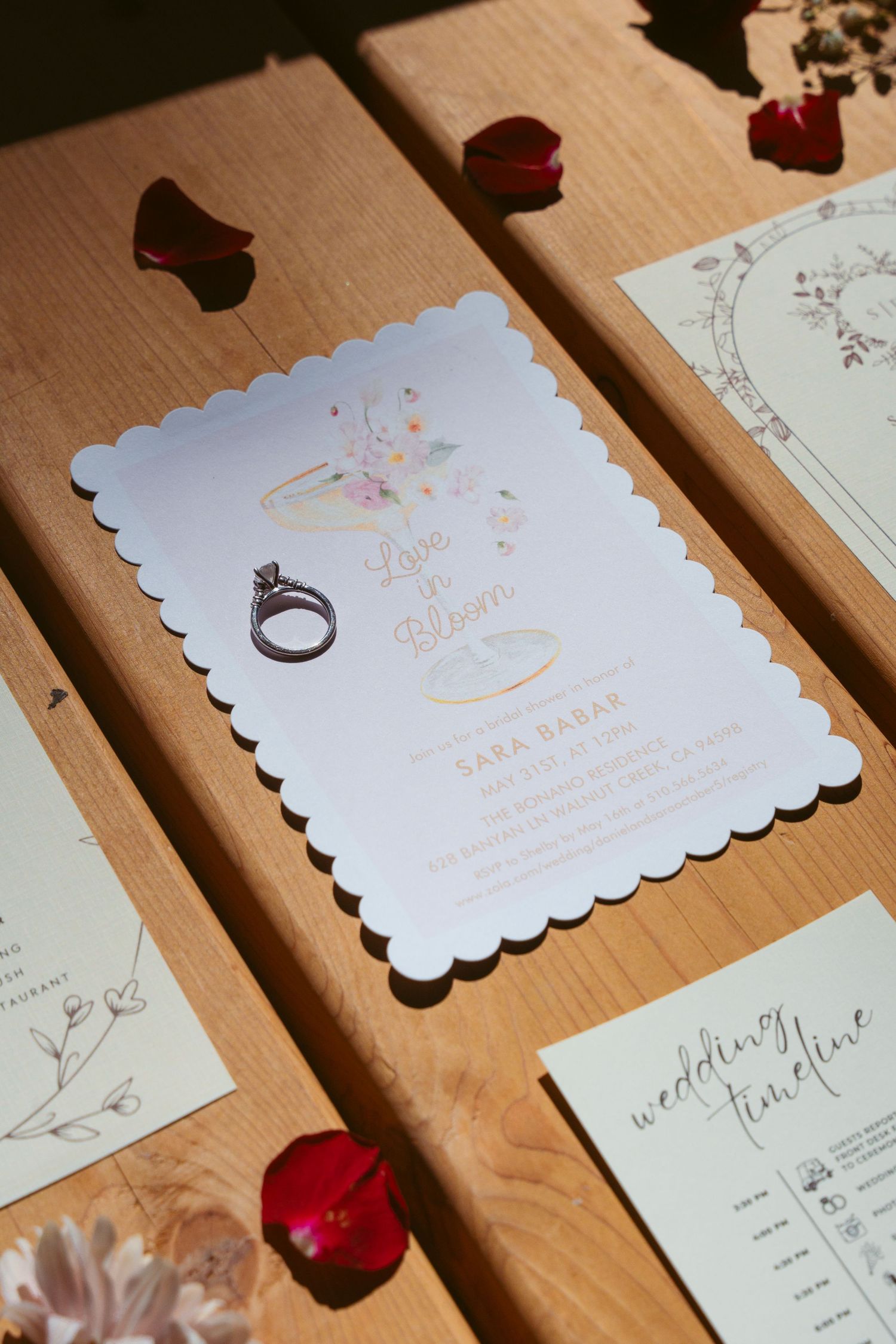 Wedding invitations and ring displayed with rose petals on wooden surface.