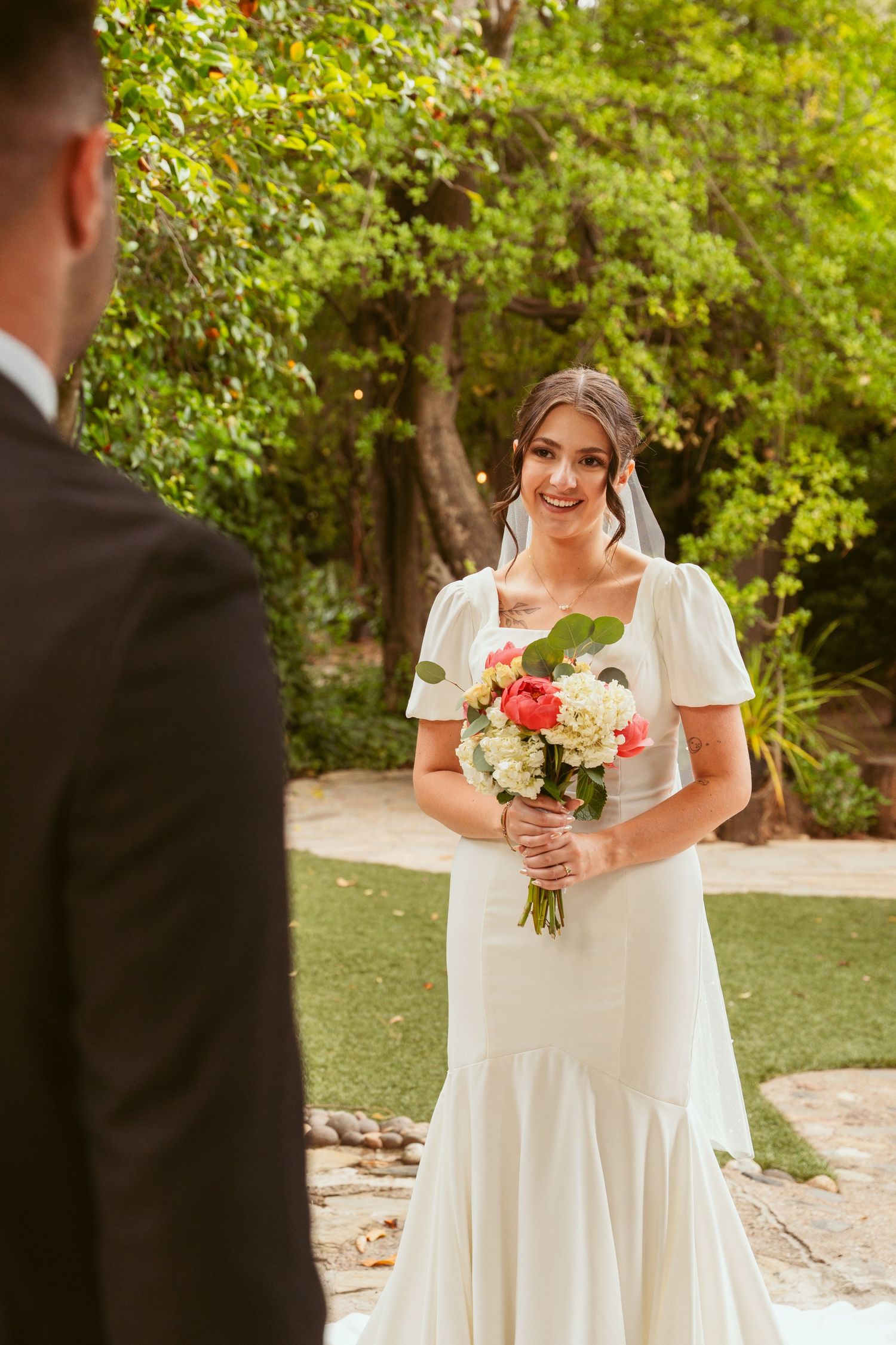 Bride in elegant white dress holds coral bouquet while looking at groom during first look.