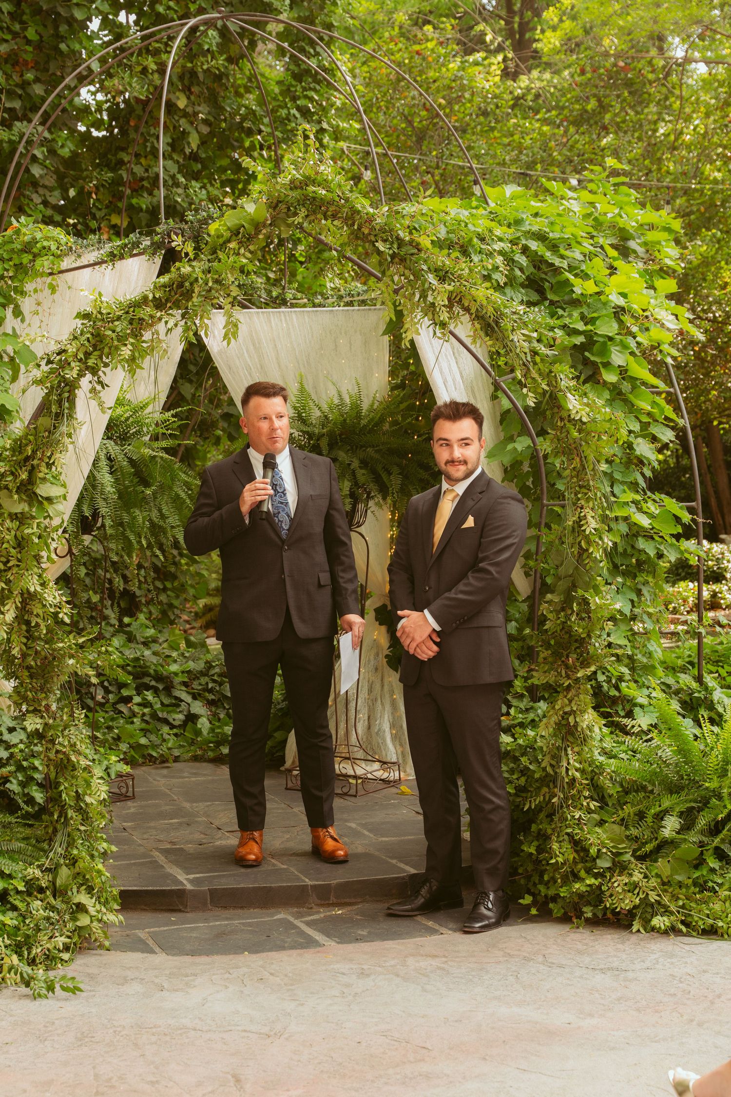 Two men in suits stand under a garden arch decorated with white fabric and greenery.