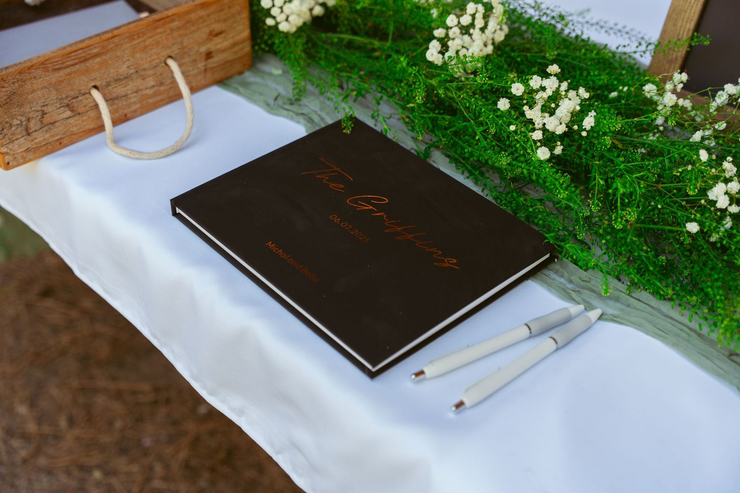 Guest book table decorated with white linens and greenery at an outdoor venue.