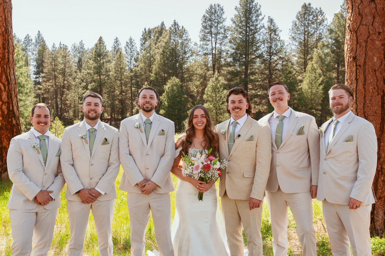 Wedding party members in light beige suits line up between rustic pine trees.