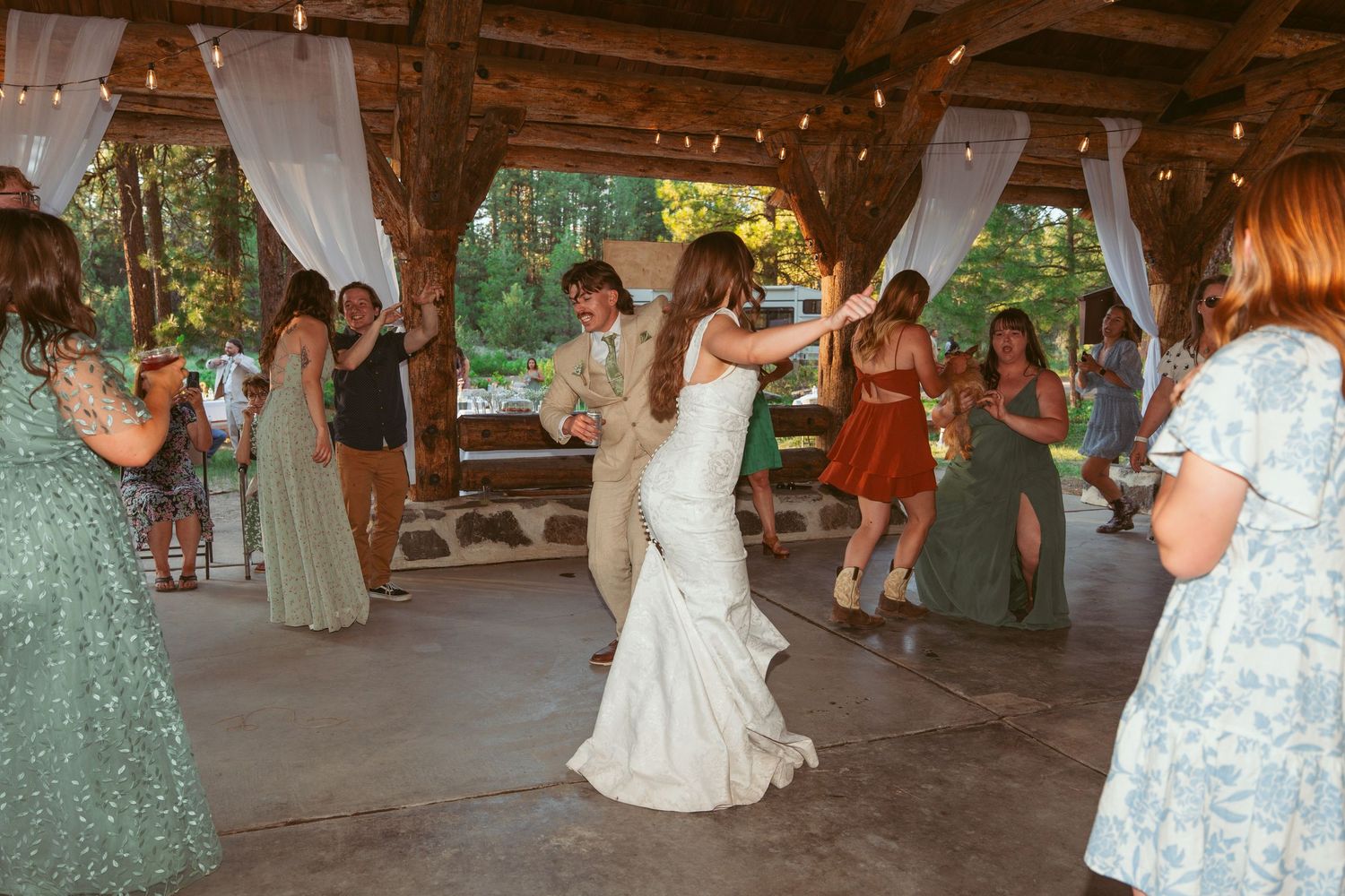 Wedding guests dance together under a rustic wooden pavilion decorated with string lights.