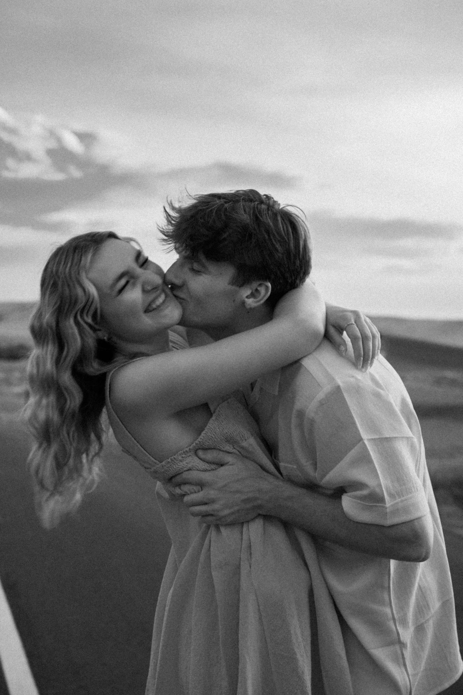 Black and white image of a couple sharing a romantic embrace against a cloudy sky.