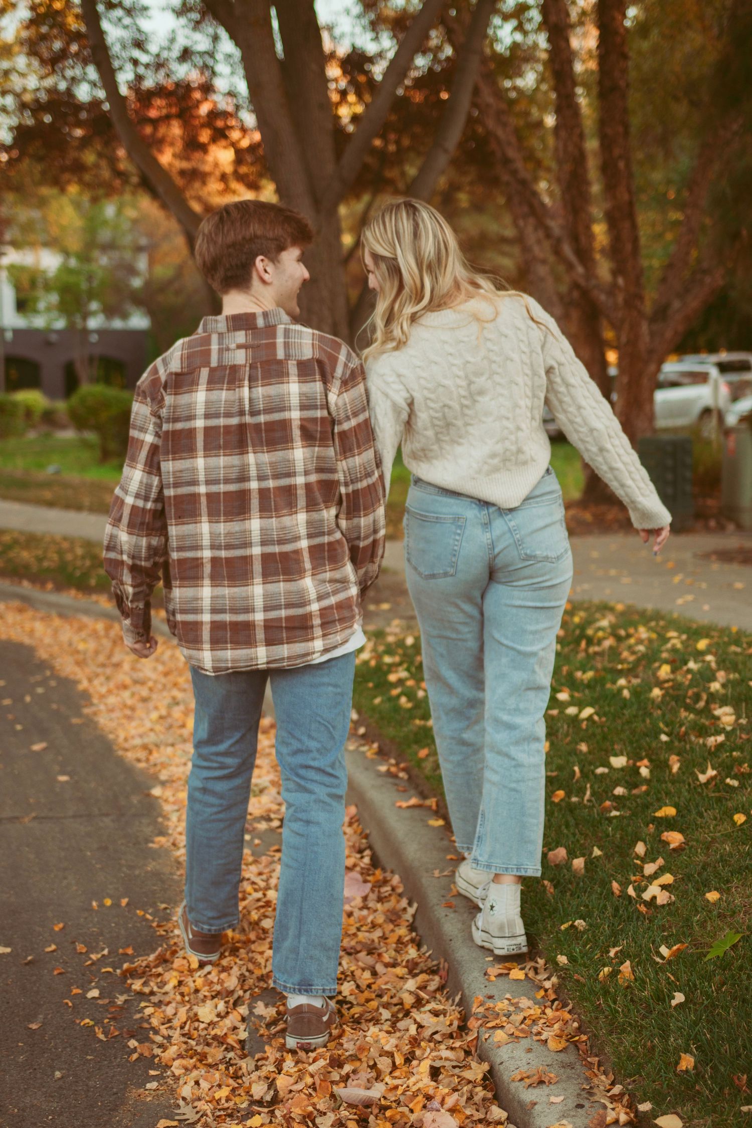 Couple walking through fallen autumn leaves on park pathway at sunset.