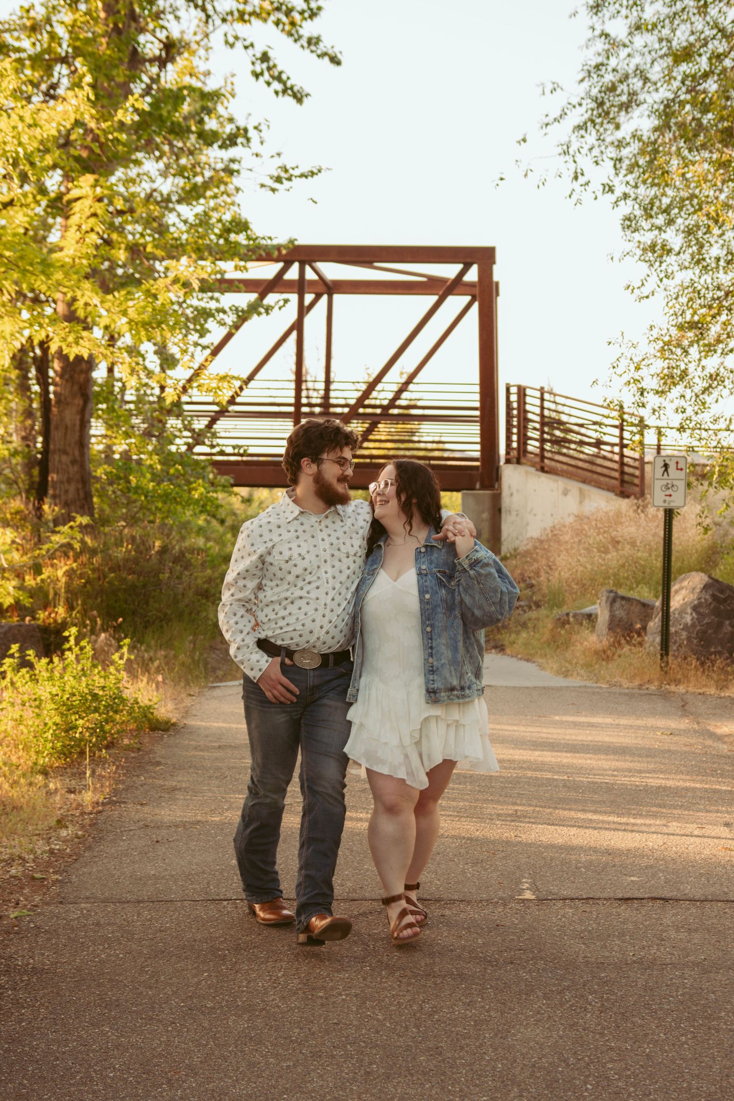 Couple walking together on a path near a metal bridge at sunset.