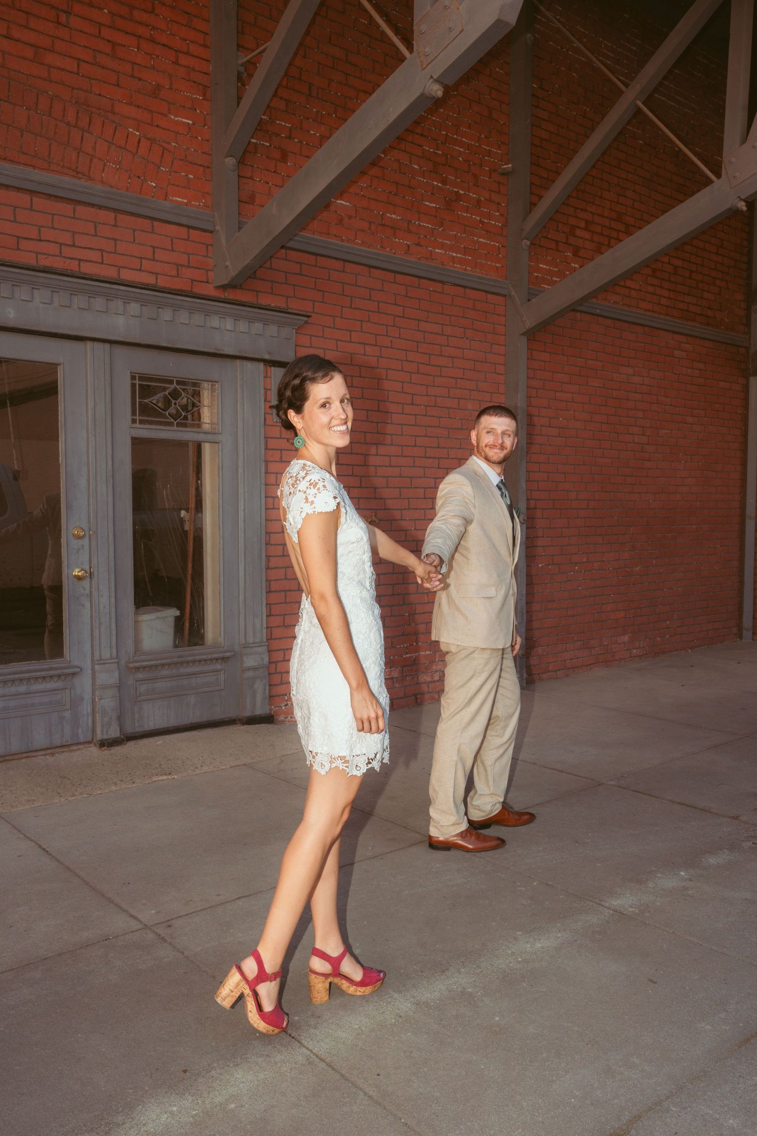 Couple in wedding attire walking playfully through brick building interior.