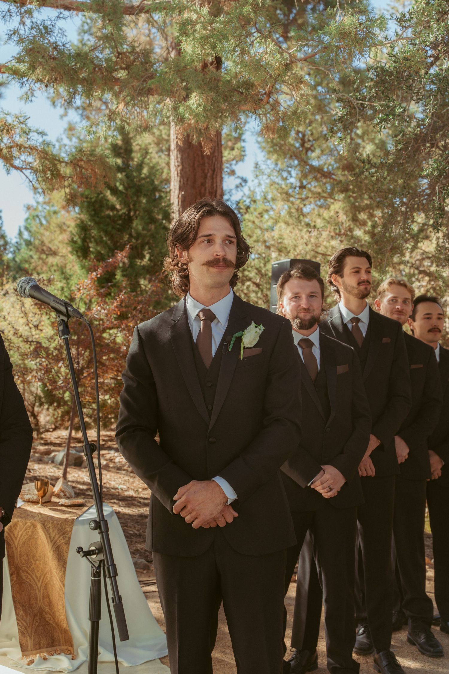 Wedding party members lined up outdoors wearing matching black suits against a natural forest backdrop.