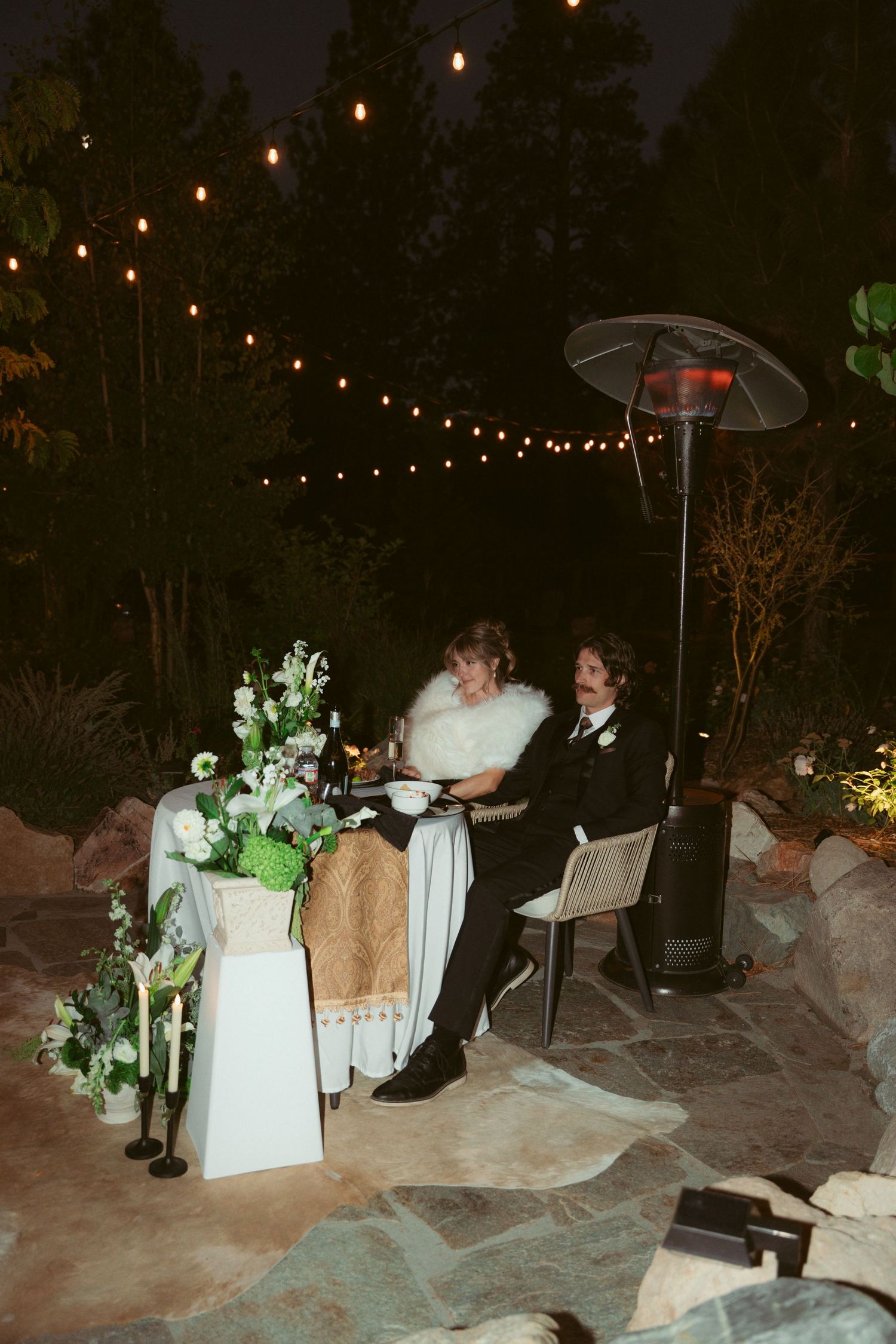 Newlyweds enjoy their wedding reception outdoors under romantic string lights and white floral arrangements.
