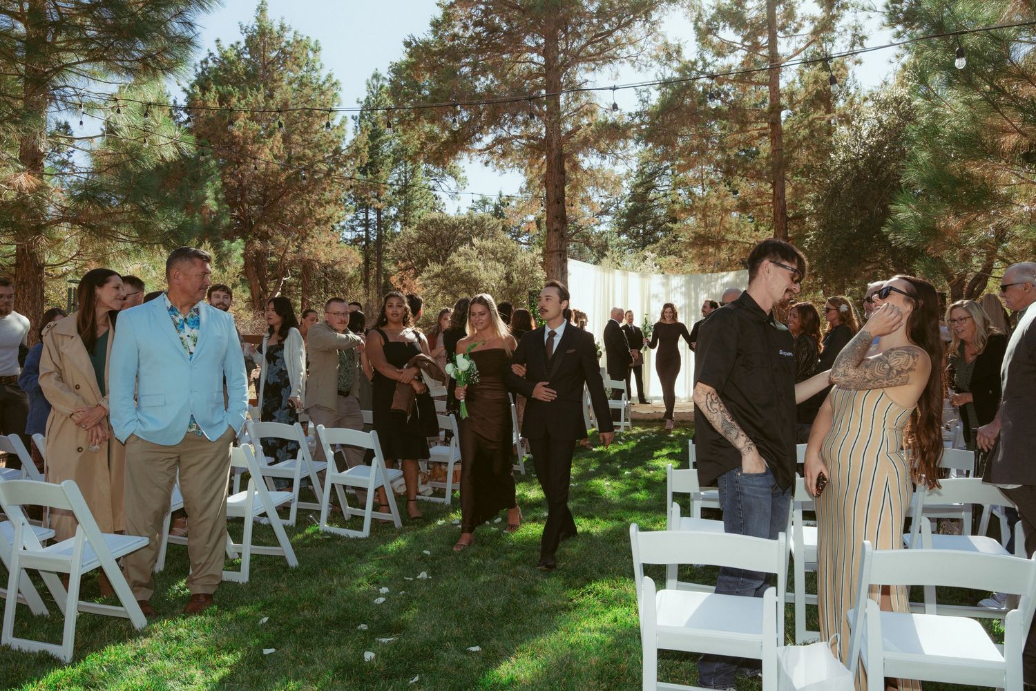 Wedding attendees socializing at outdoor ceremony venue with white chairs and pine trees.