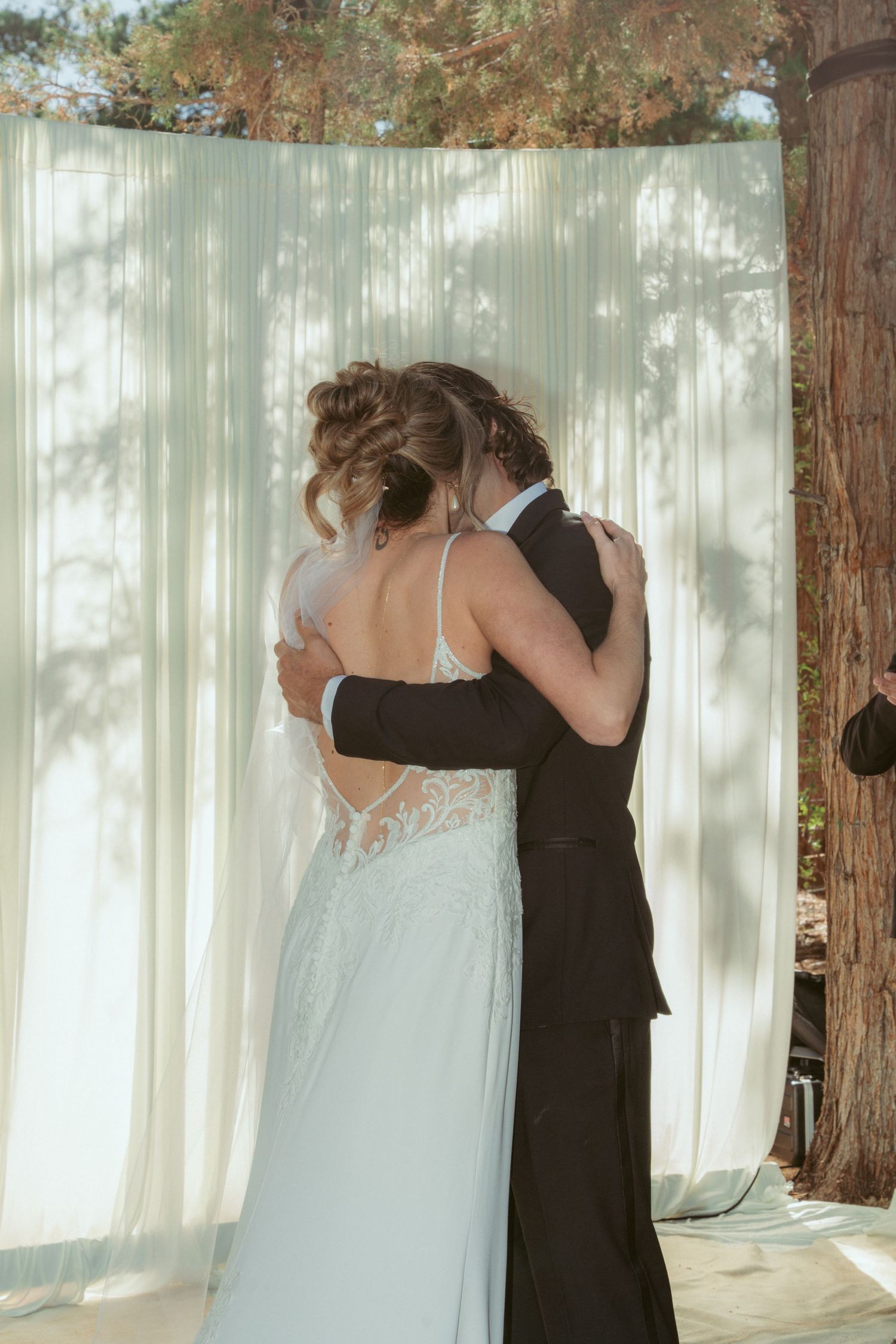 Couple embracing during wedding ceremony against white curtain backdrop in forest setting.