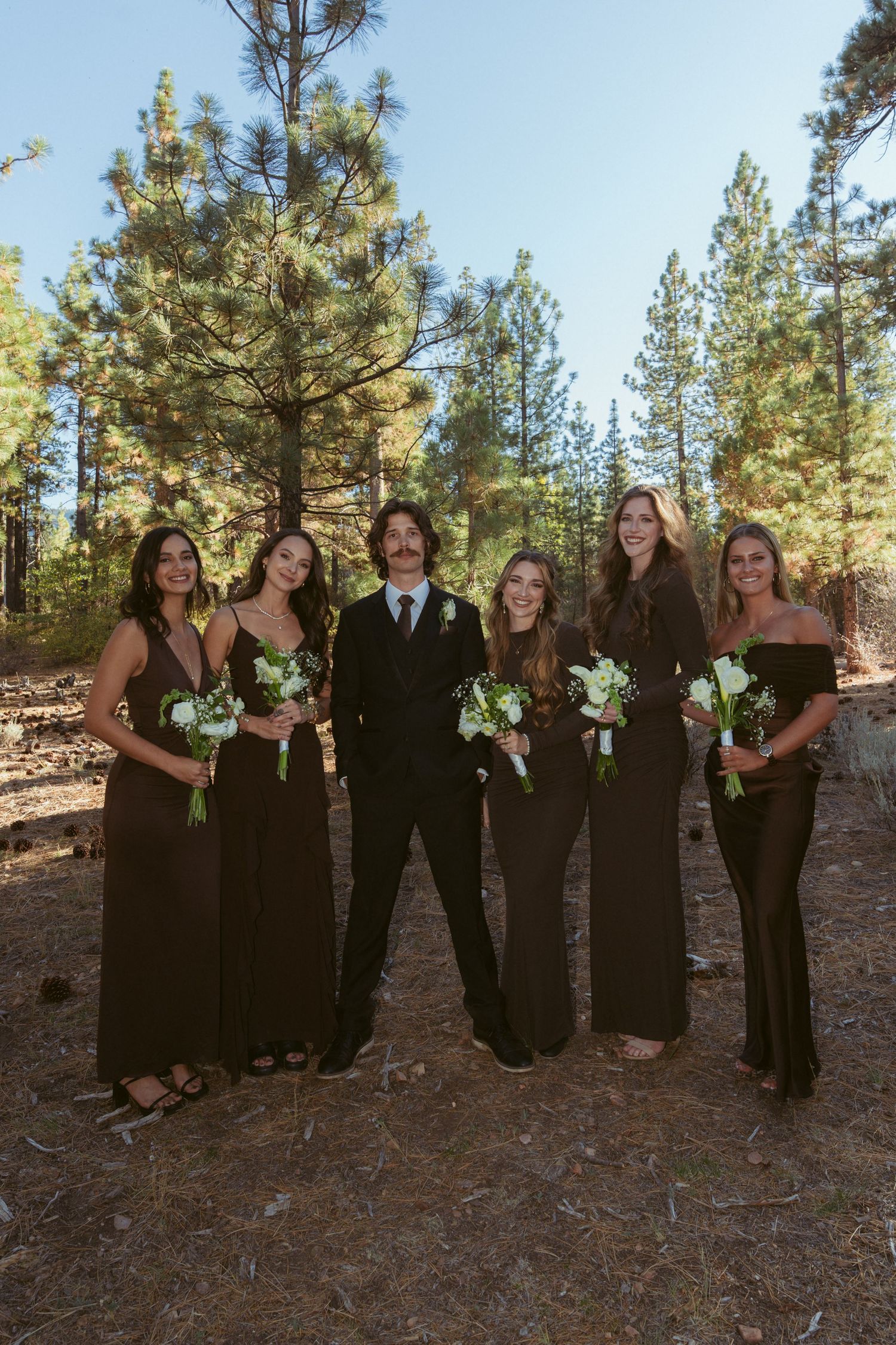 Wedding party poses in forest setting wearing black dresses and suits with green bouquets.