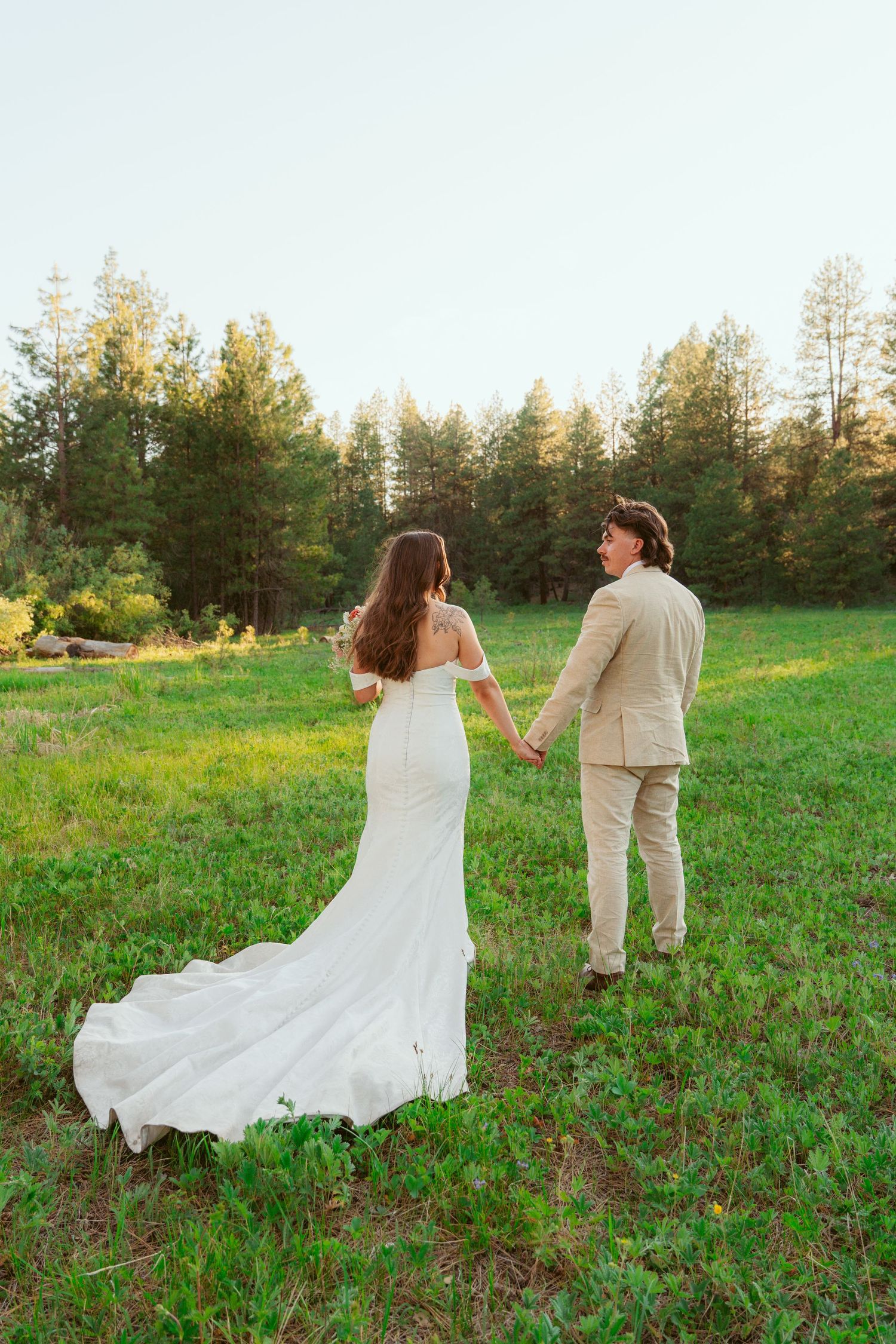 Couple in wedding attire walking and dancing through a green field near forest.