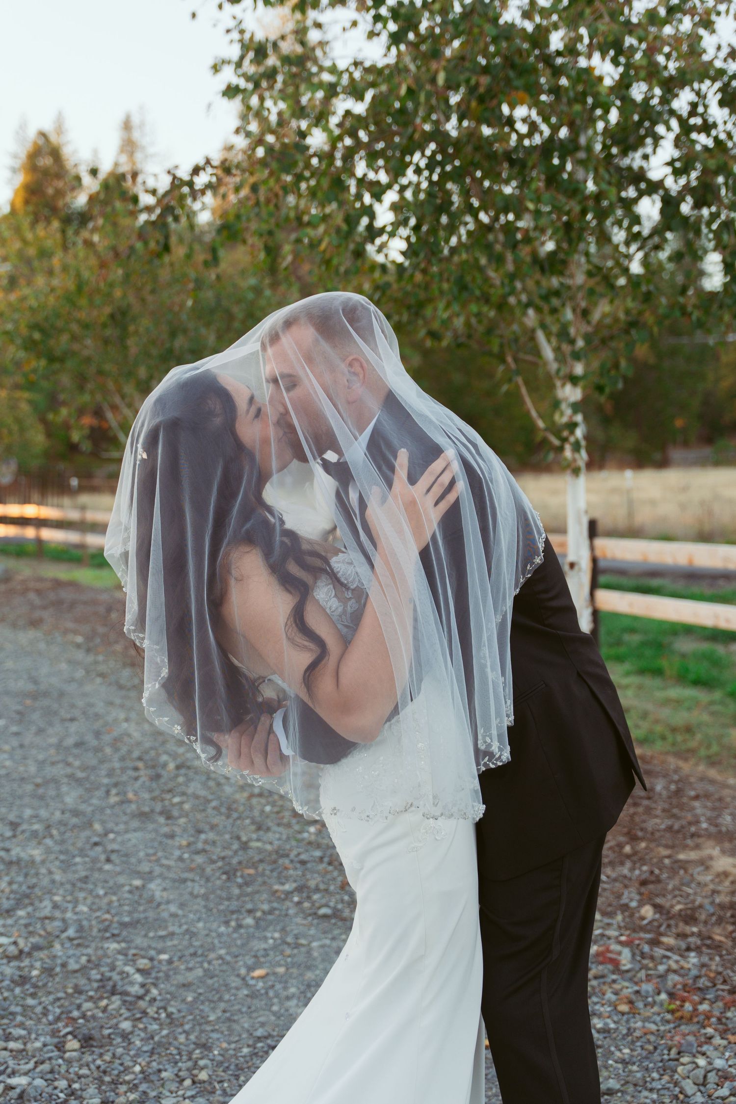 Couple shares romantic kiss under a delicate white veil in an outdoor setting.