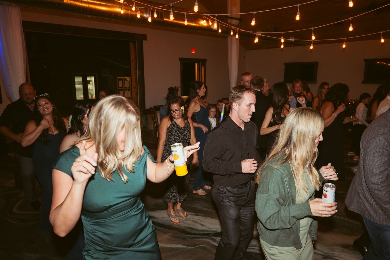 Wedding guests dance and celebrate on a dimly lit dance floor under string lights.