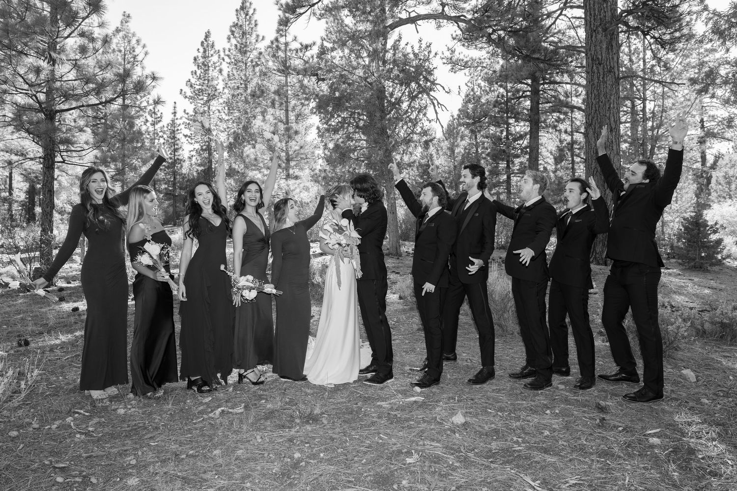 Black and white photo of wedding party celebrating with raised arms in forest setting.