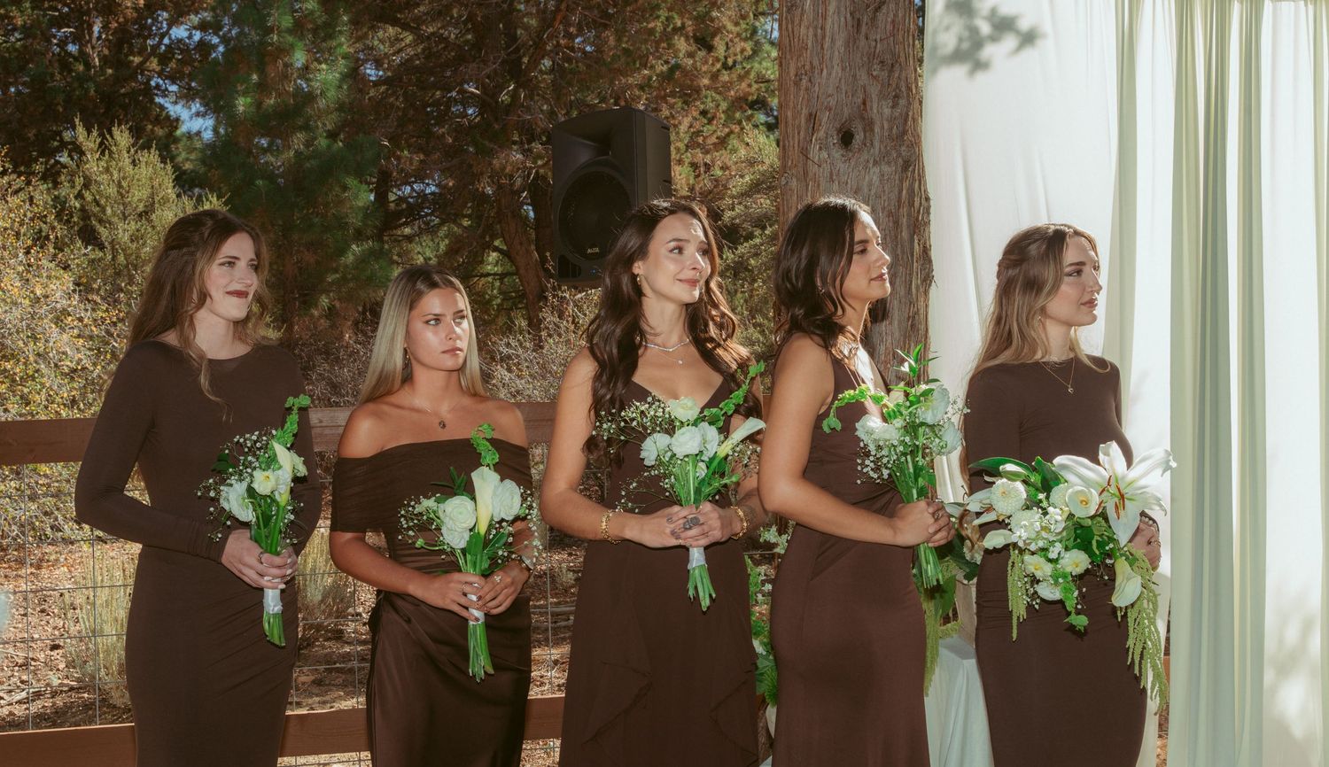 Wedding party in brown dresses holding green and white bouquets during outdoor ceremony.