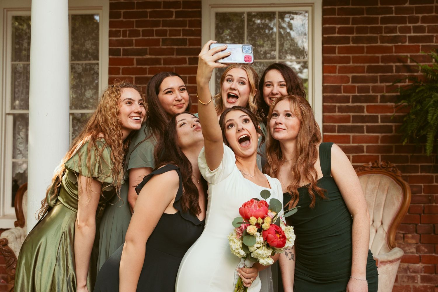 Bridesmaids in green dresses take selfie together while laughing and holding bouquet.