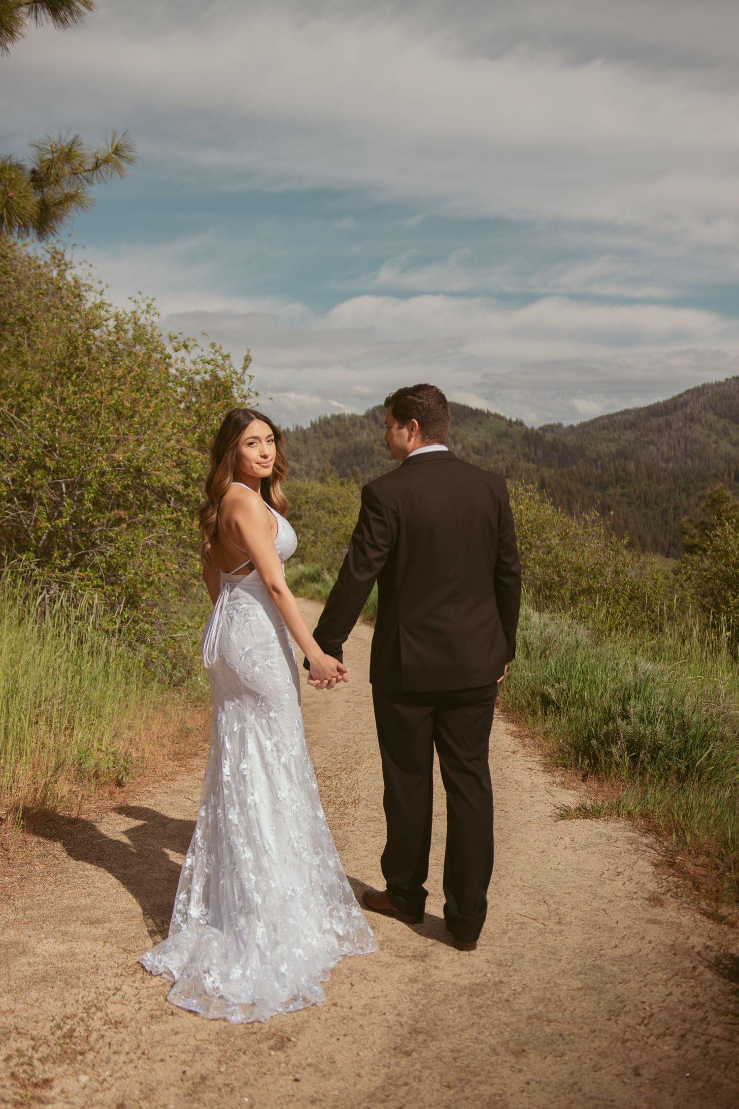 A couple in wedding attire hold hands while walking on a mountain trail surrounded by nature.