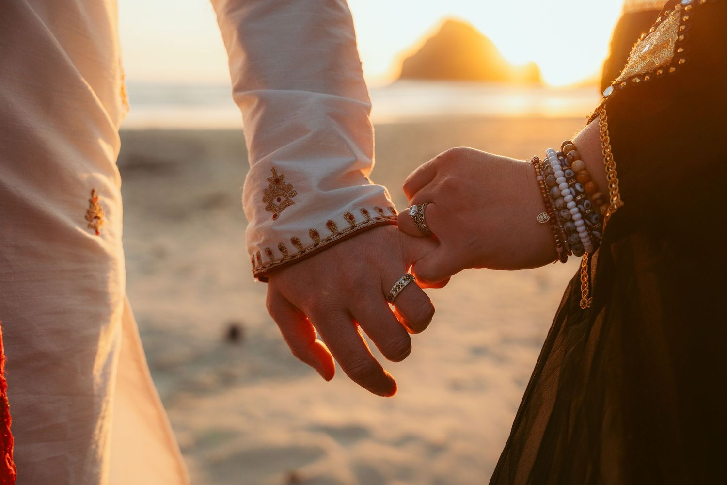 Close-up of hands with ornate bracelets touching during a sunset beach ceremony.