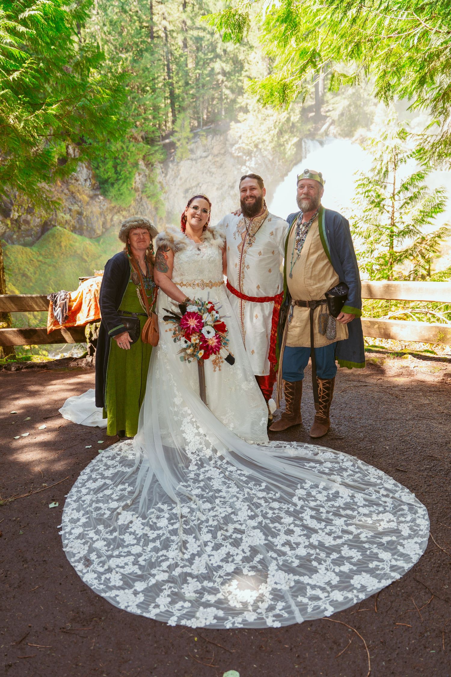 A wedding party poses in medieval-inspired attire against a wooded forest backdrop with a flowing white lace dress train.