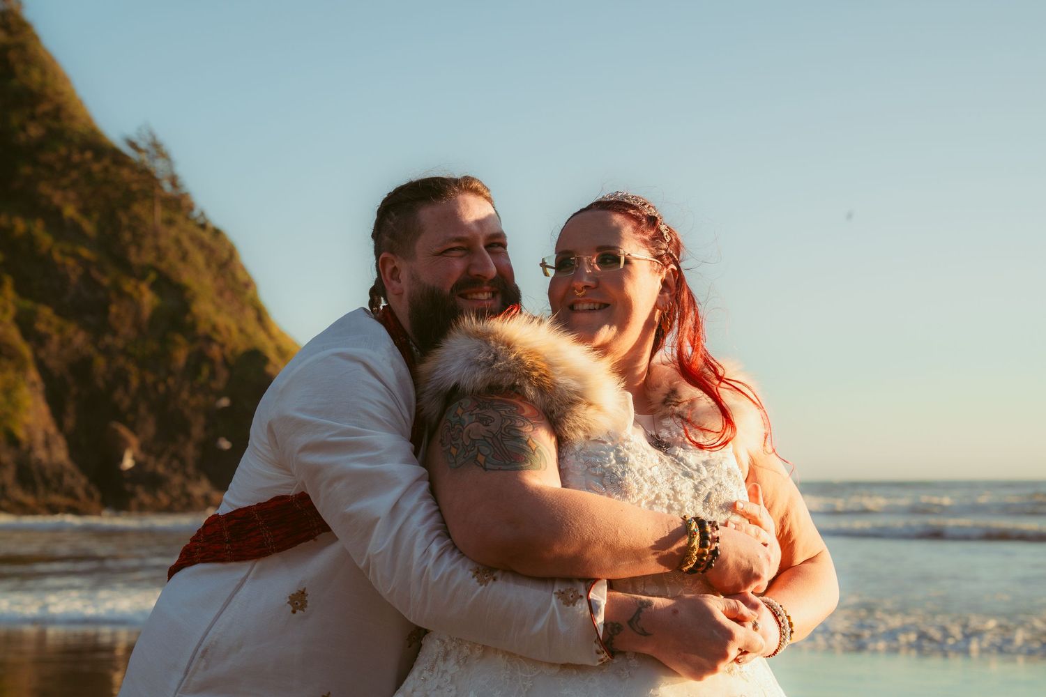 A couple in white wedding attire embraces on a scenic beach with waves crashing in the background.
