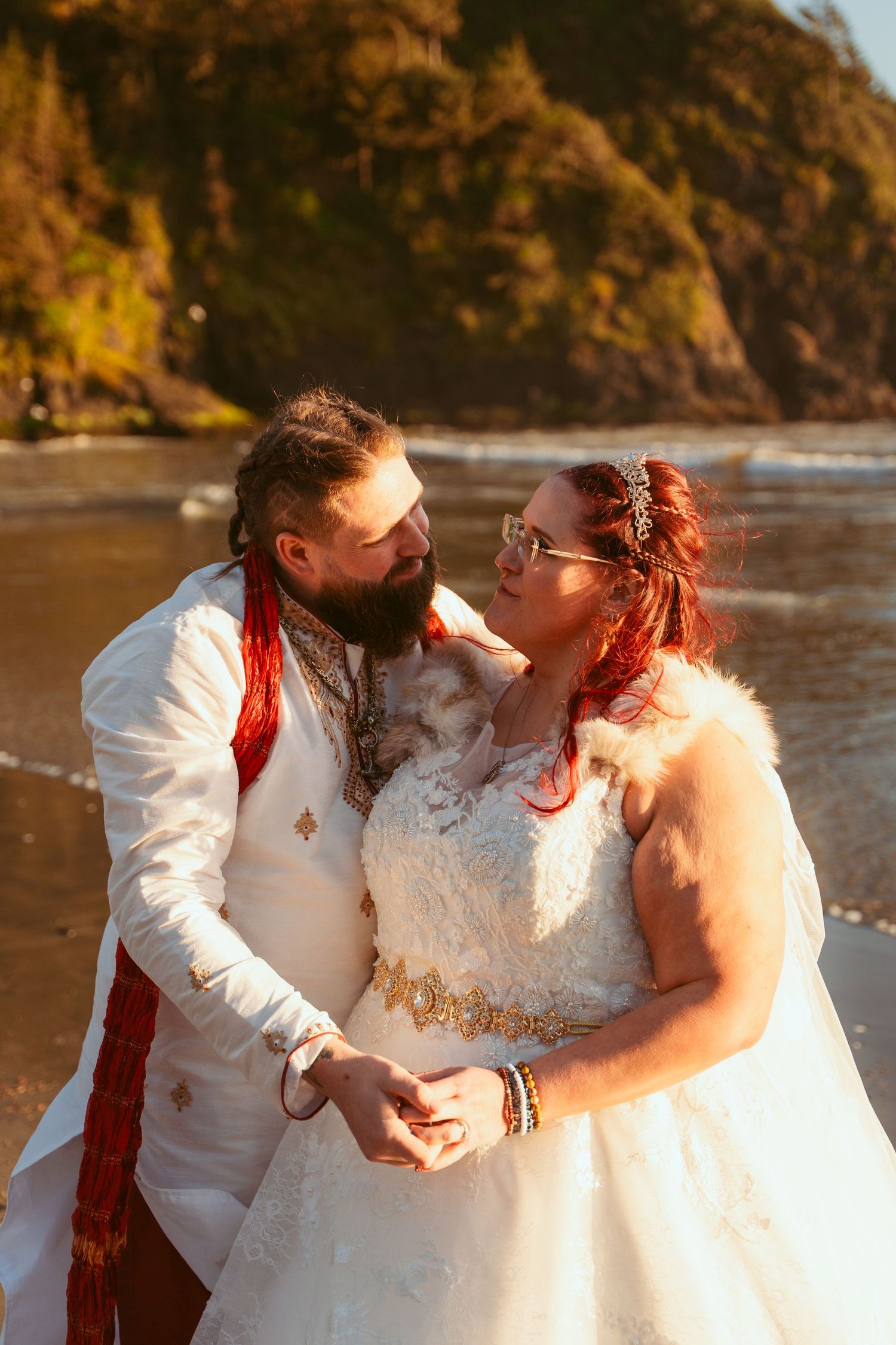 Newlyweds in white and red attire embracing on beach with cliffs in background.