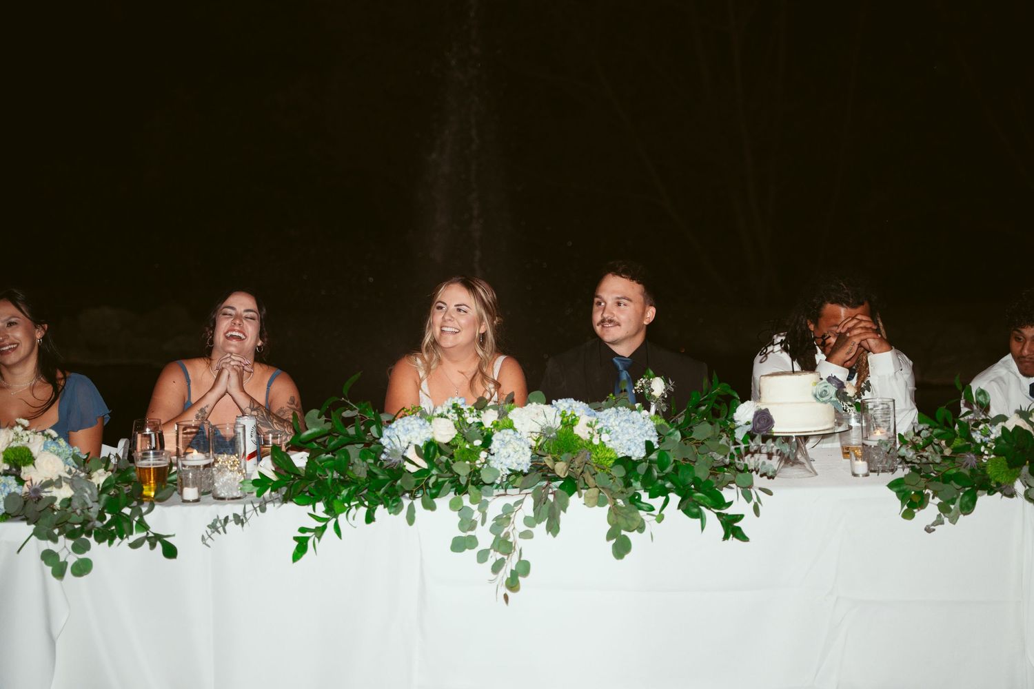 Wedding guests seated at a decorated head table with greenery and white linens laugh and applaud during celebrations.