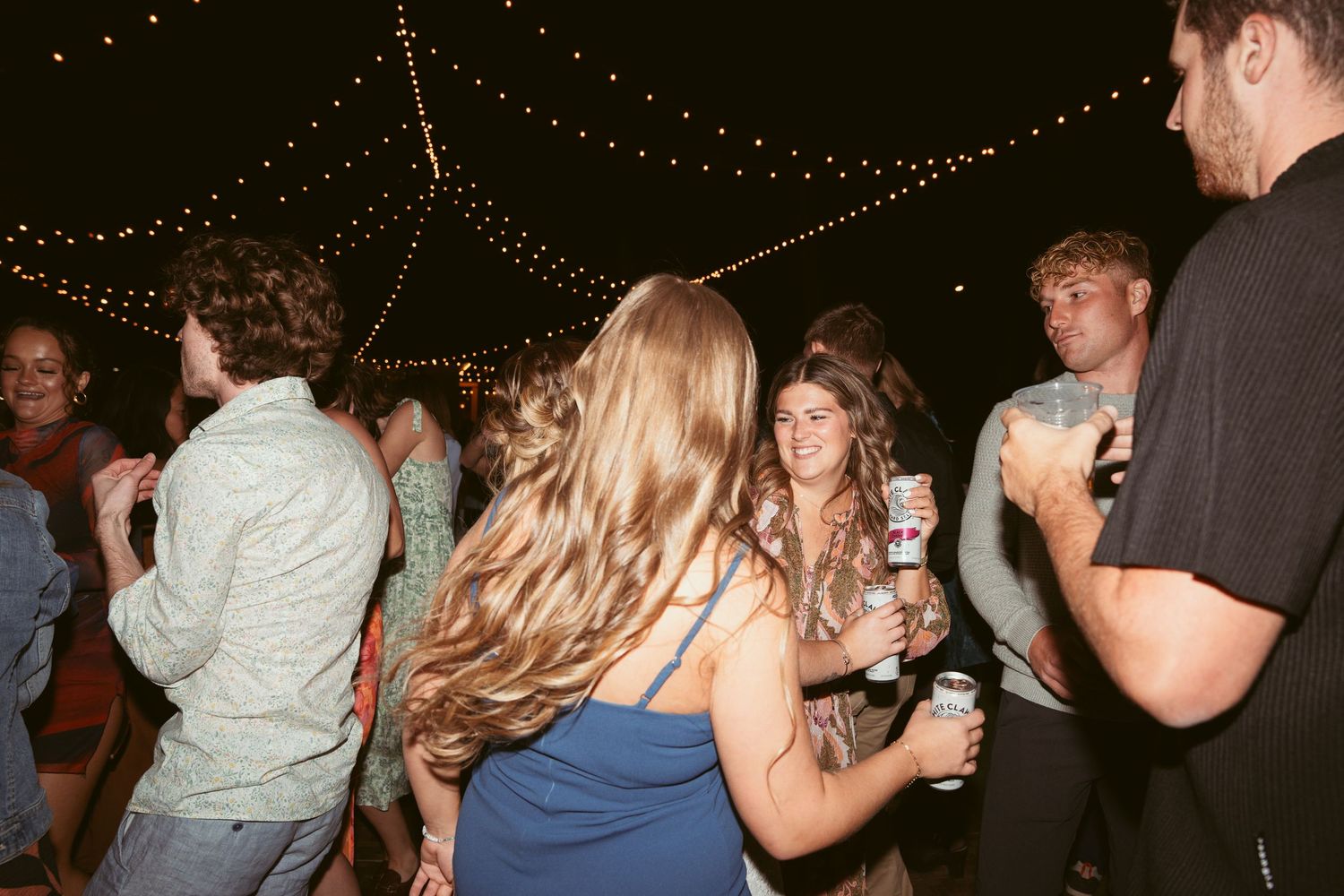 Crowd mingling and dancing at a dimly lit party with string lights overhead.