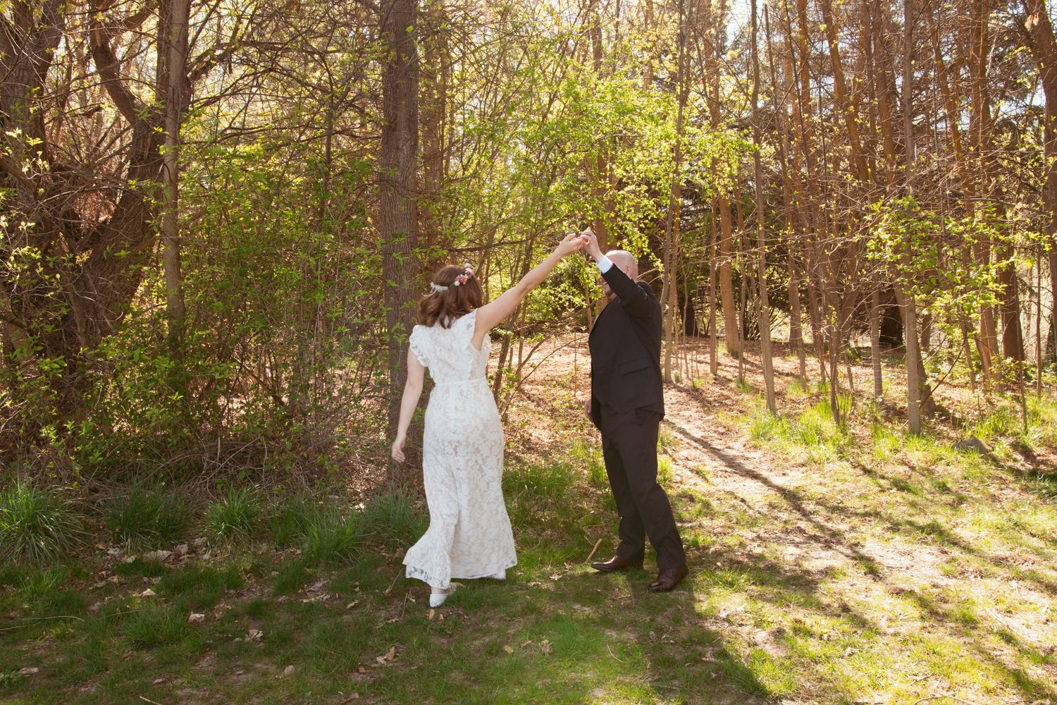Newlyweds walk hand in hand through a sunlit forest path in their wedding attire.