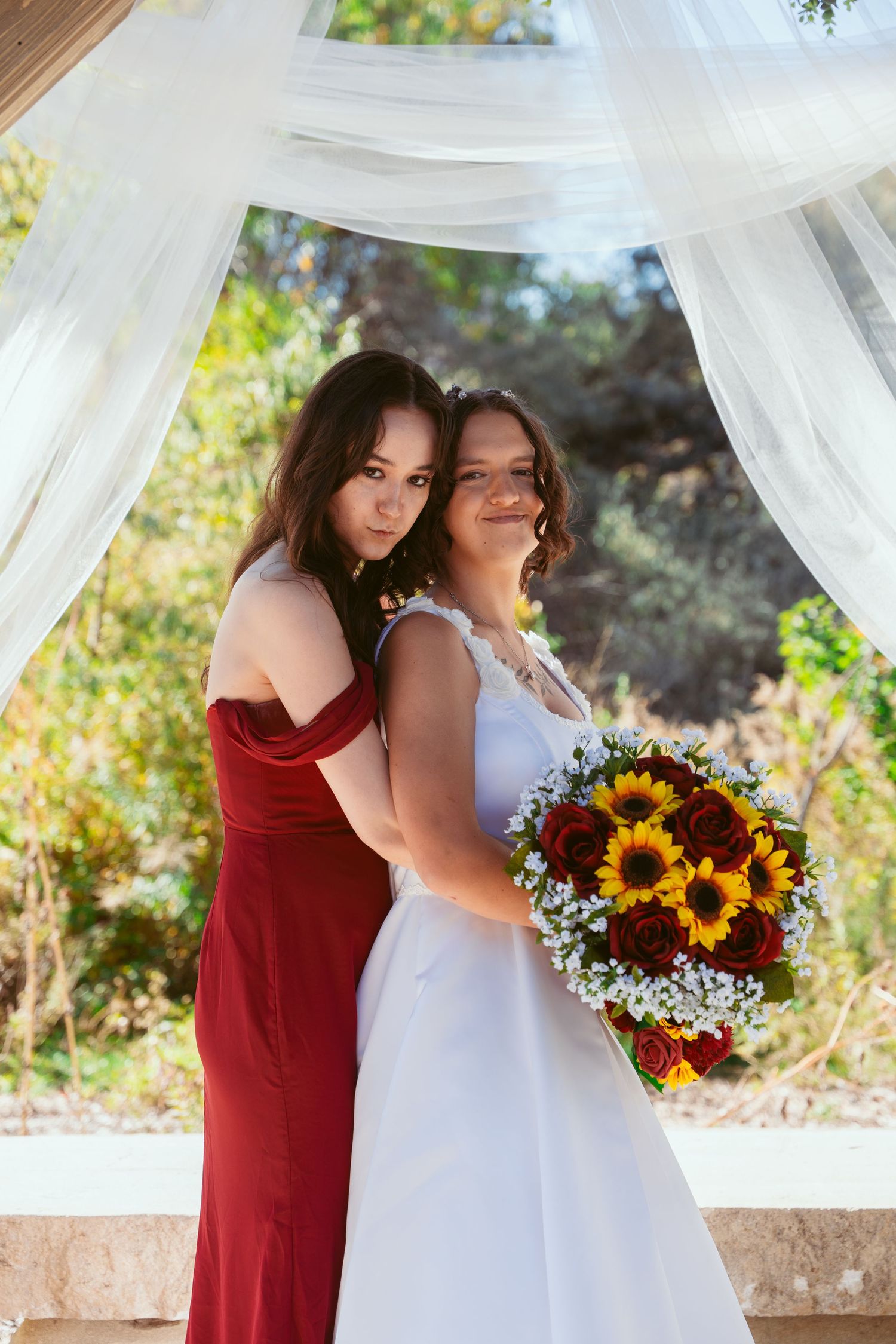 Wedding couple embraces under white curtains with vibrant sunflower and red rose bouquet against outdoor backdrop.