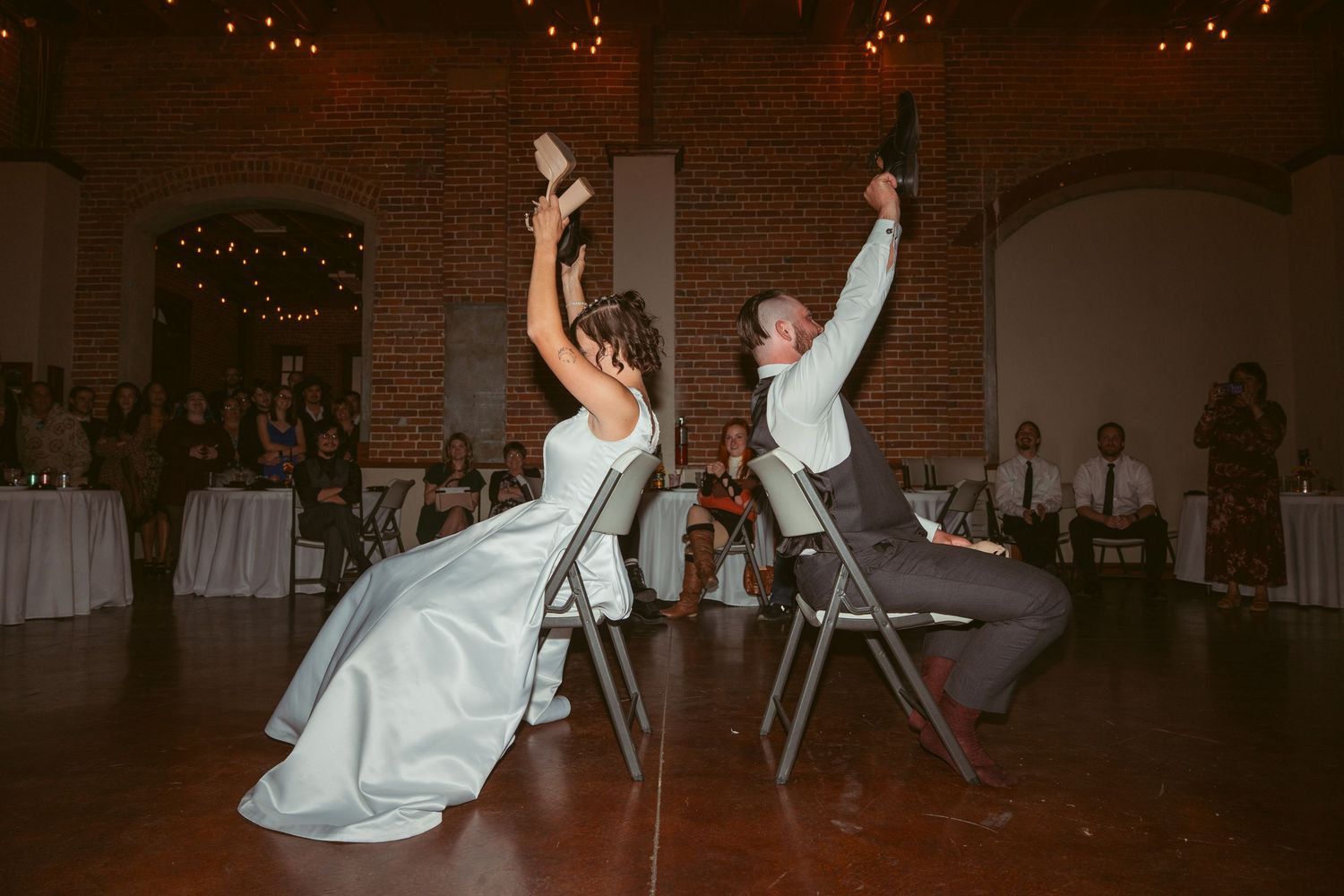 Wedding party members dance in formation at an indoor reception with exposed brick walls.