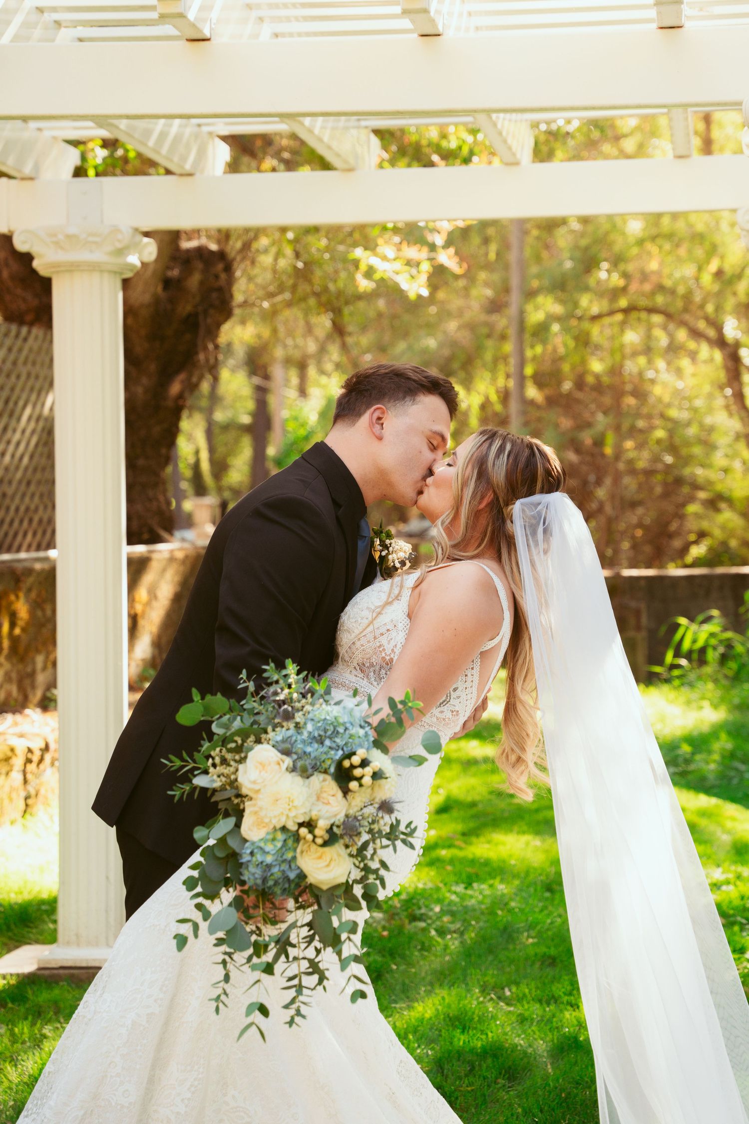 A romantic wedding moment captured under a white pergola with flowing bouquet and veil details.
