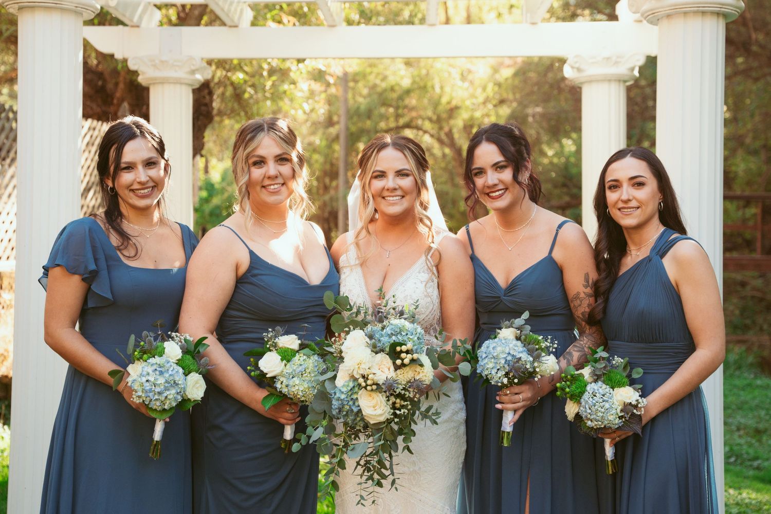 Wedding party in navy blue dresses poses with bouquets under white garden archway.