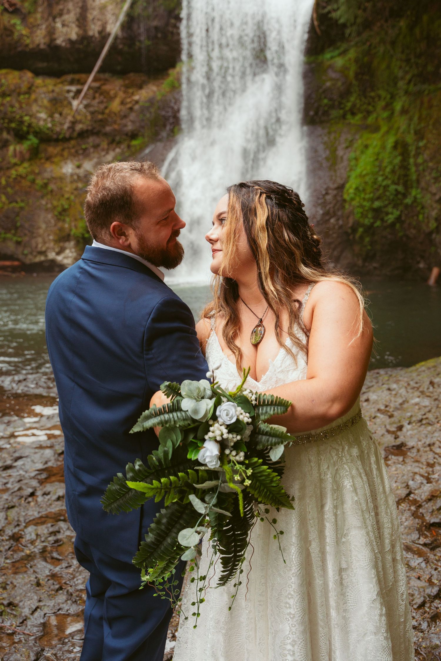 Wedding couple embraces while holding a white and green bouquet near a forest waterfall.