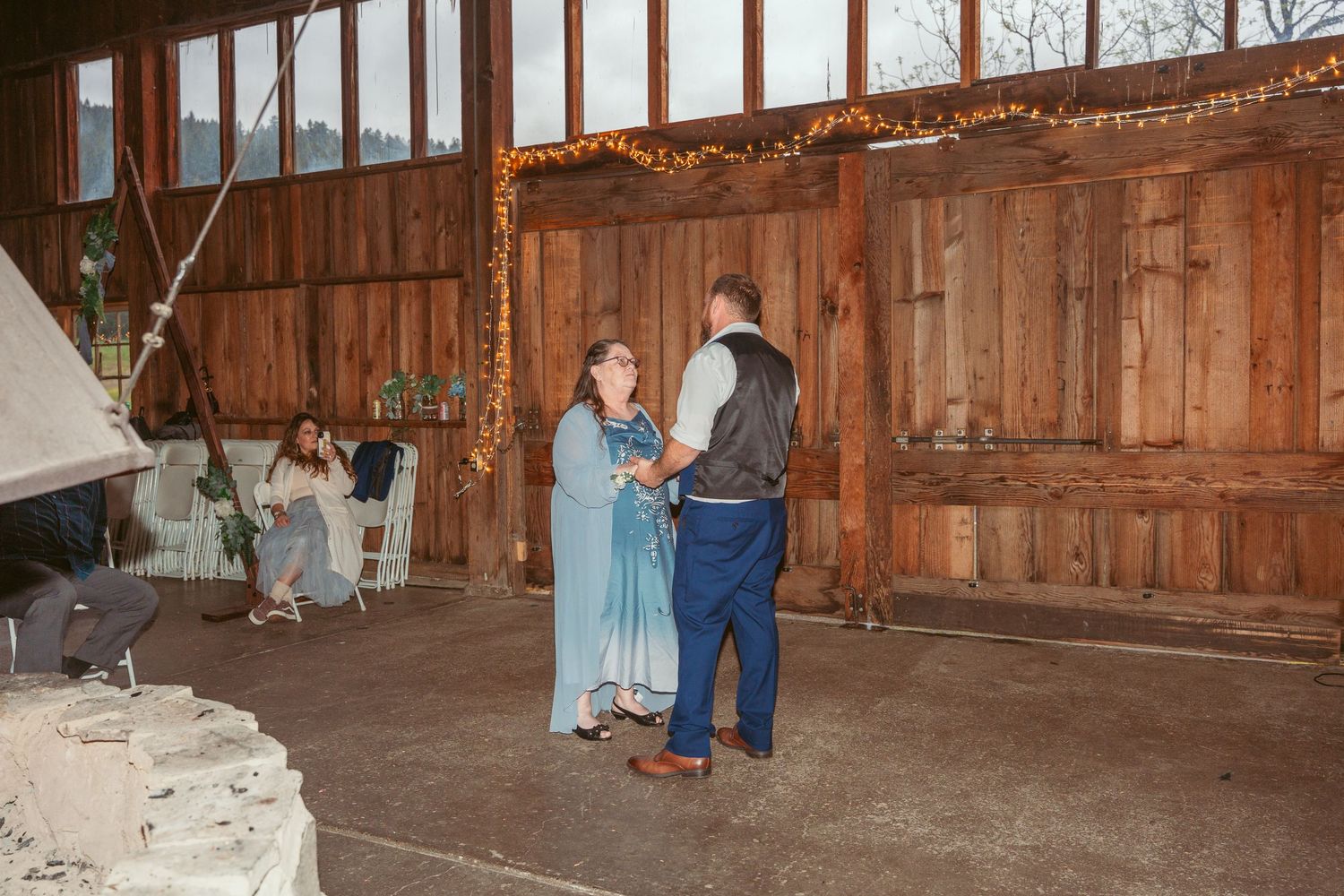 People dancing in a spacious rustic barn venue with large windows and string light decorations.