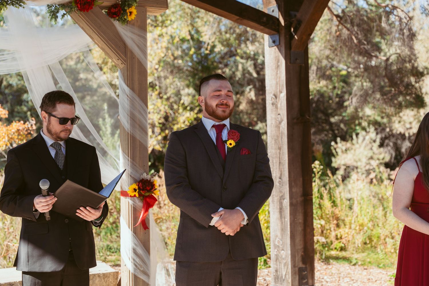 Wedding officiant and groomsmen standing under wooden pergola decorated with autumn flowers.