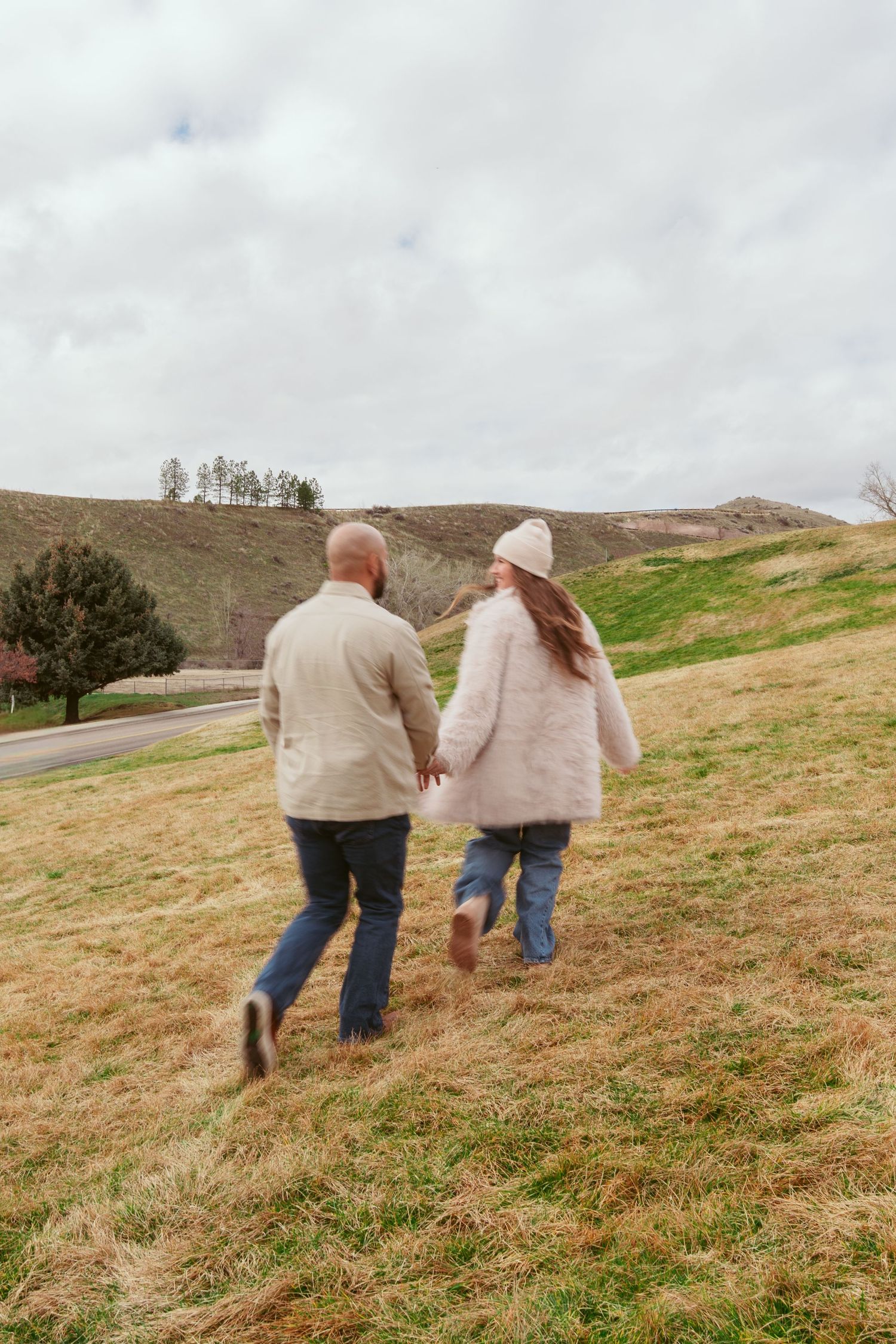 A couple runs hand in hand up a grassy hillside on a cloudy winter day.