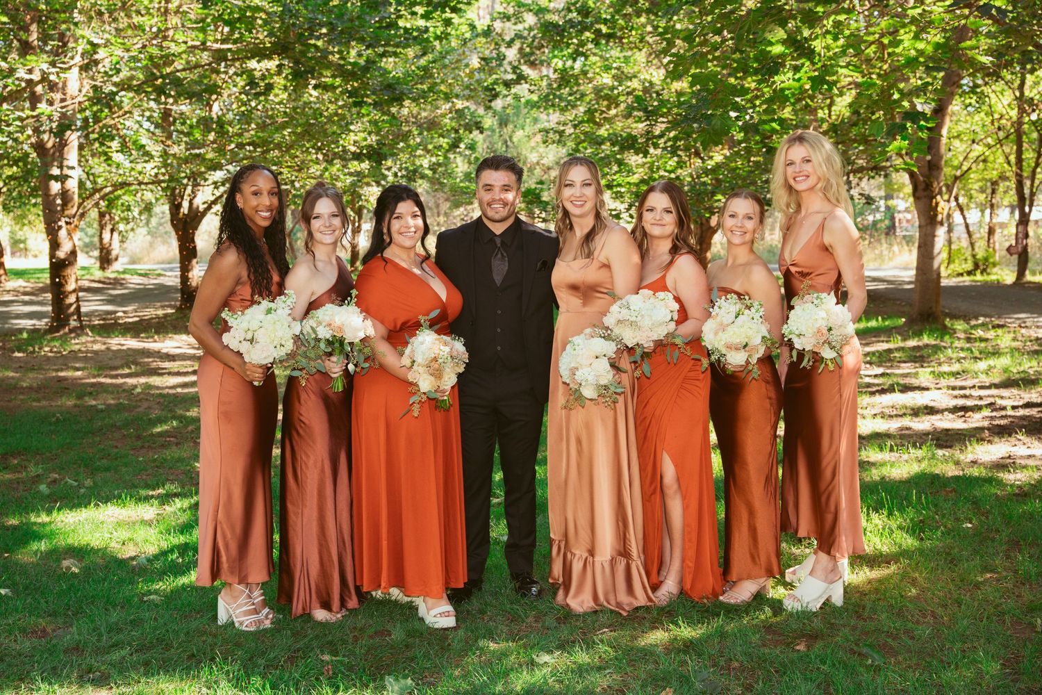 Wedding party in coordinated coral and peach dresses poses together under green trees in a park setting.