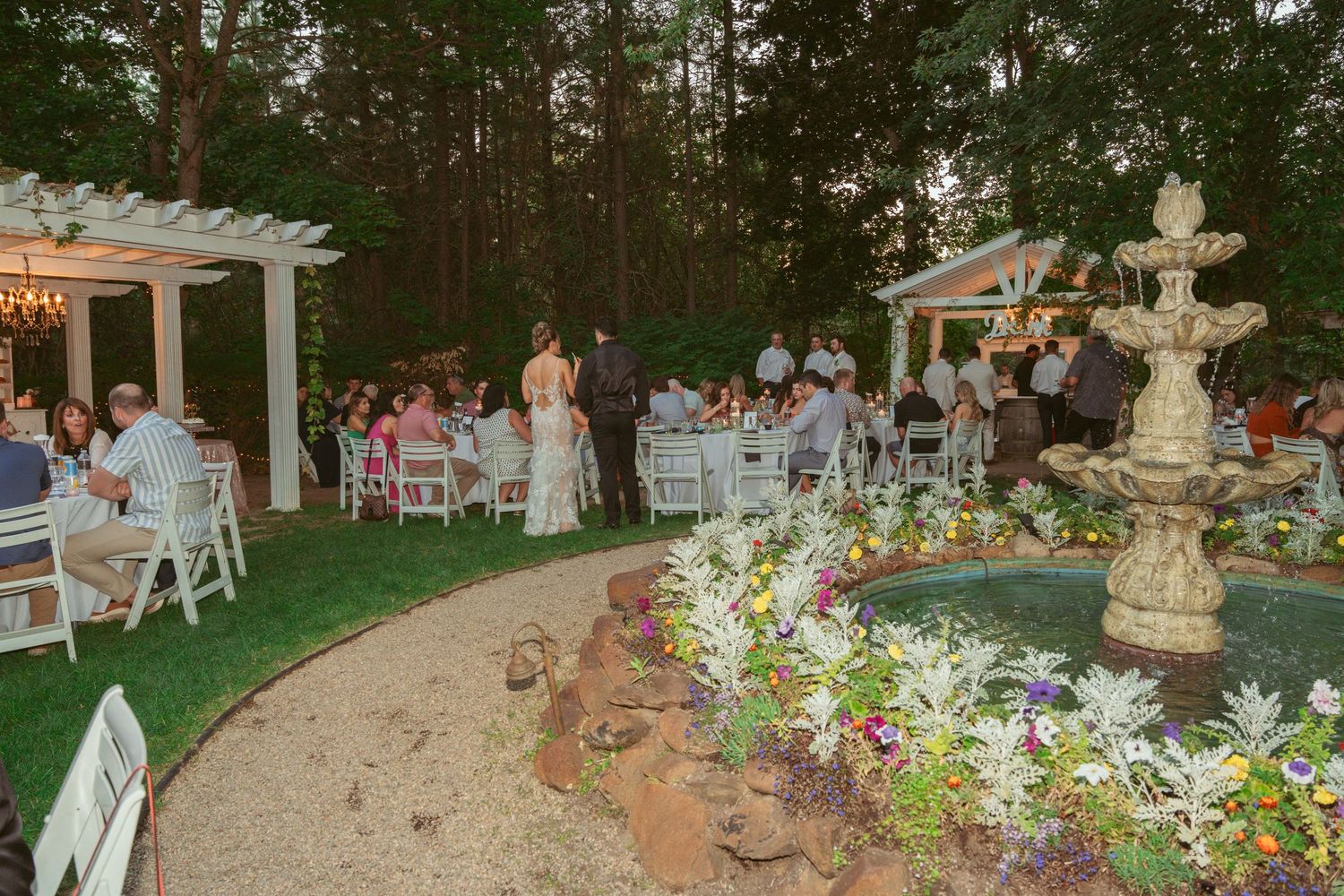 Outdoor wedding reception setup with white chairs and tables arranged under string lights in a garden setting.