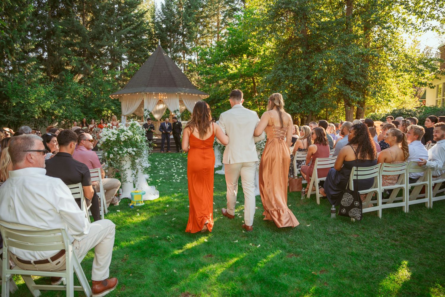 Wedding guests in orange and white attire gather on a sunlit lawn for an outdoor ceremony.