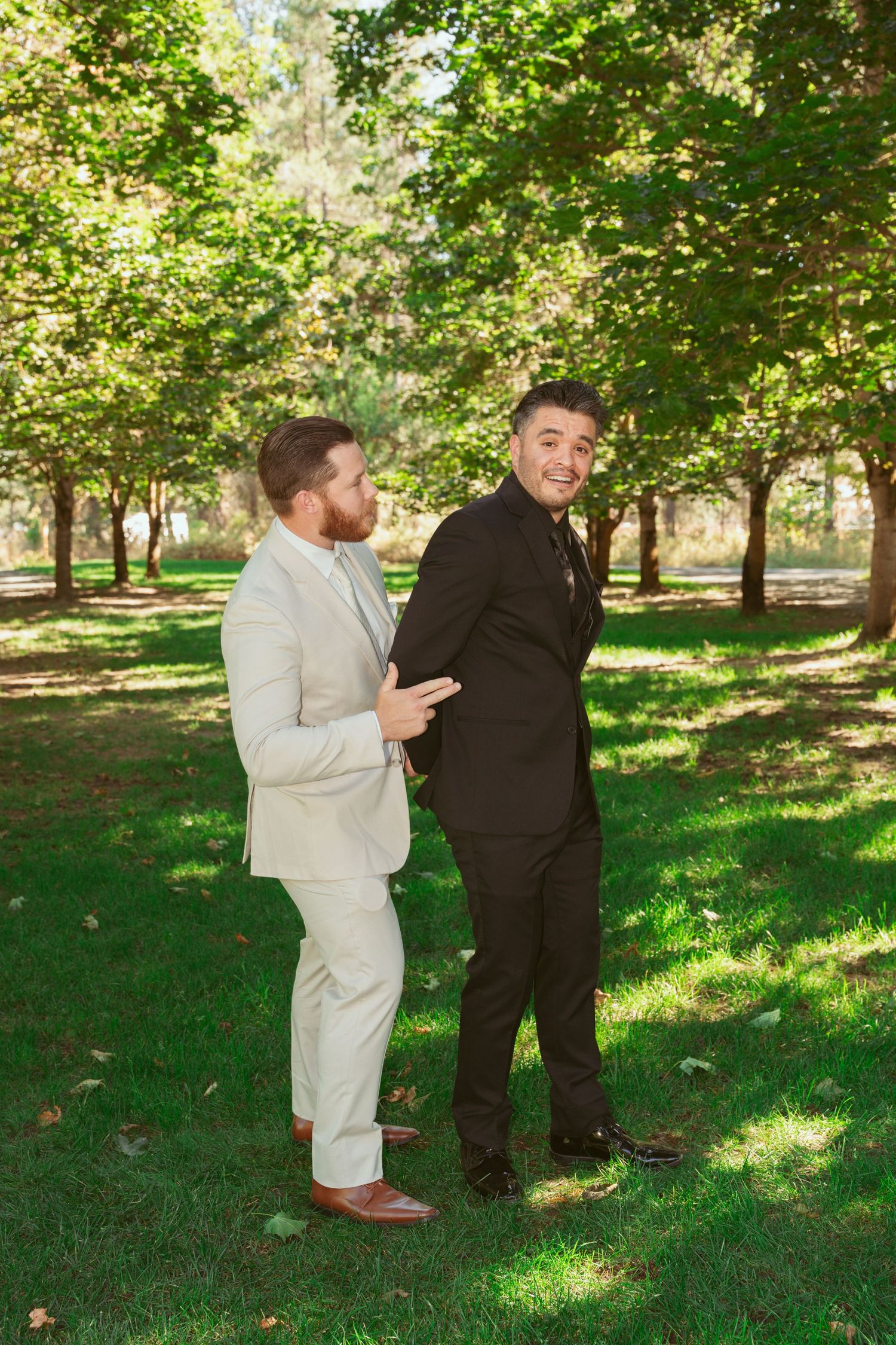 Two people in contrasting light and dark formal attire share a candid moment on grassy lawn under trees.