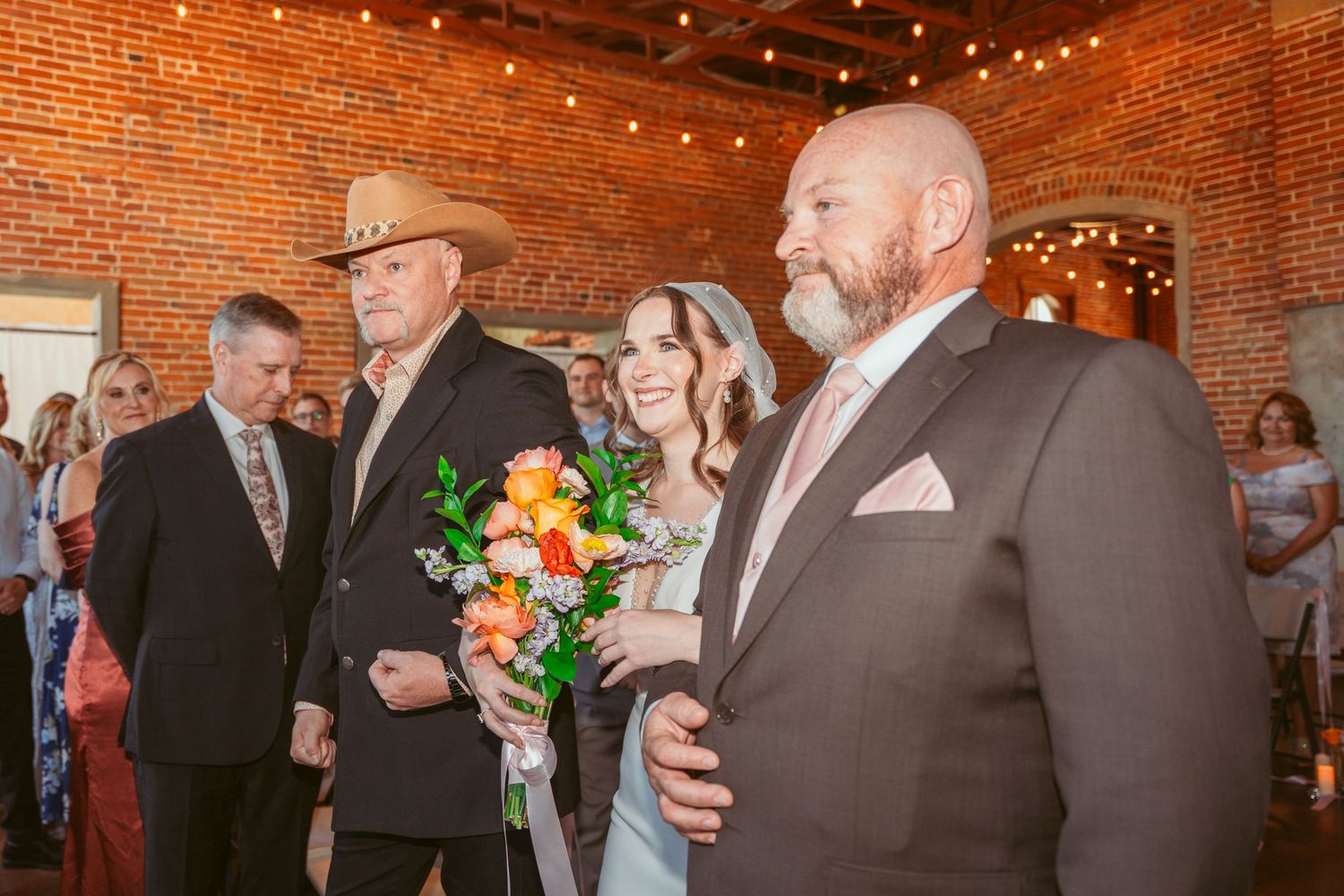 Wedding guests gather in a rustic brick venue decorated with string lights during a reception celebration.